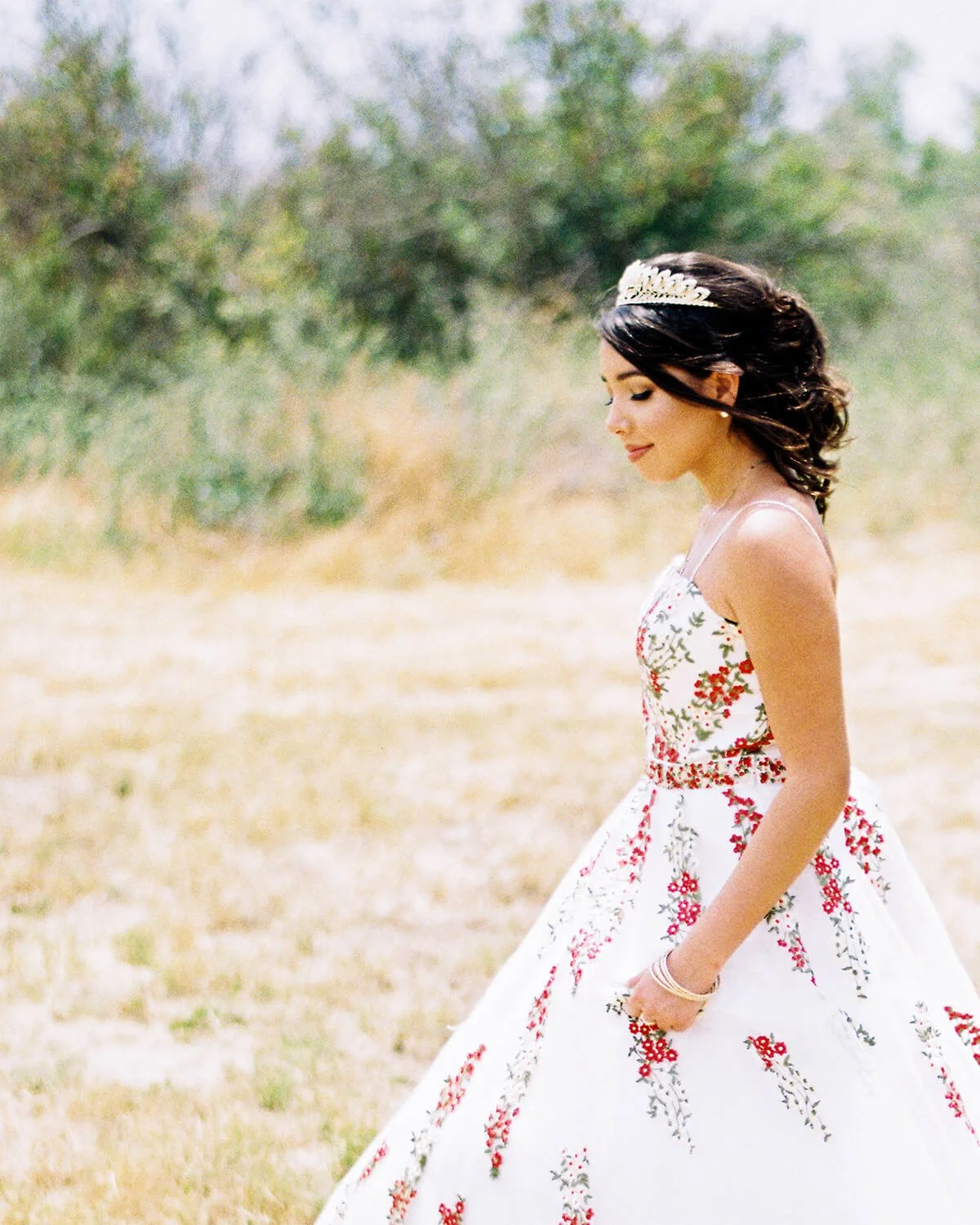 A Quinceañera in a floral wedding dress with a tiara, standing outdoors in a grassy area with trees in the background.