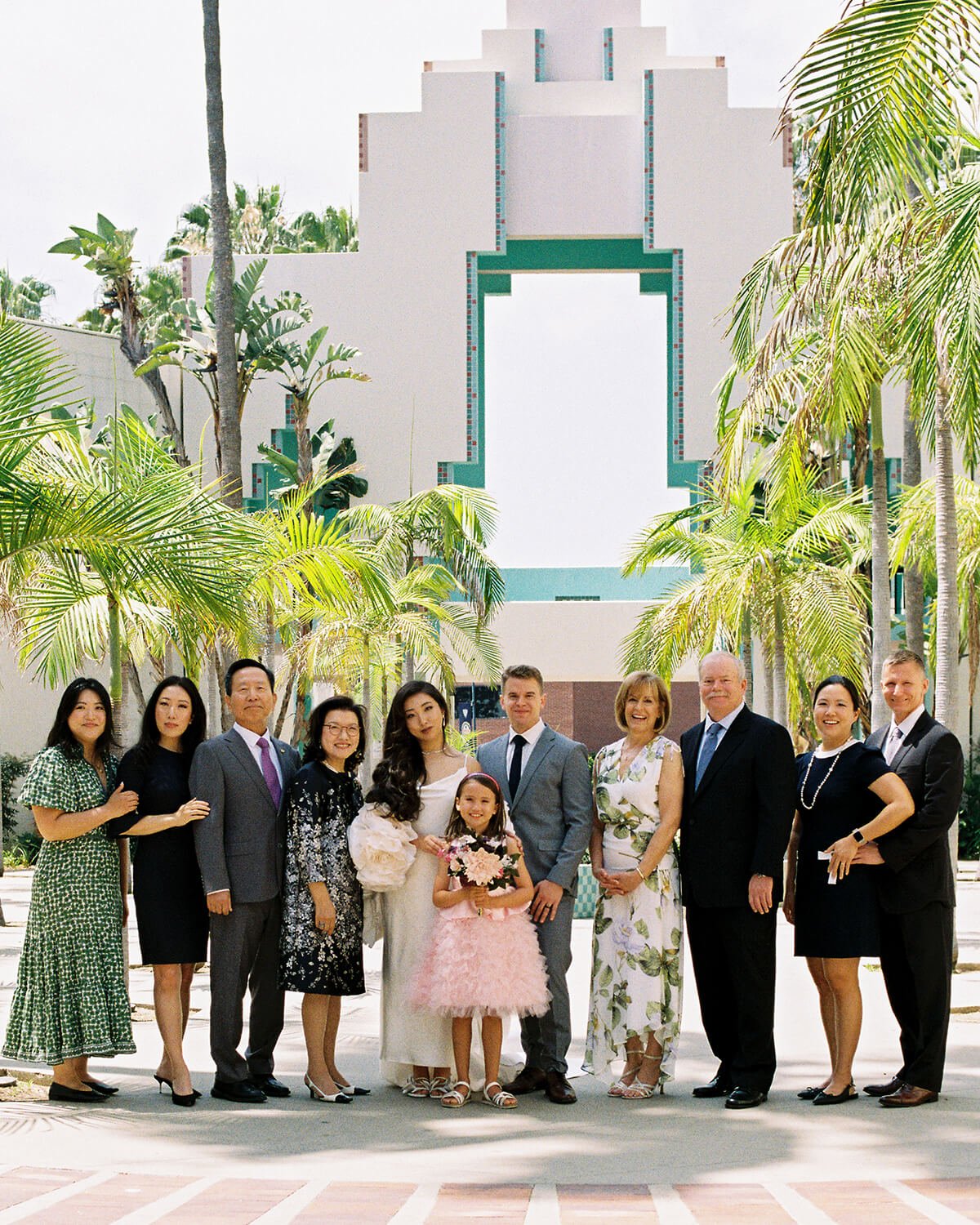 Group of people in formal attire standing outdoors in front of a white building with palm trees.