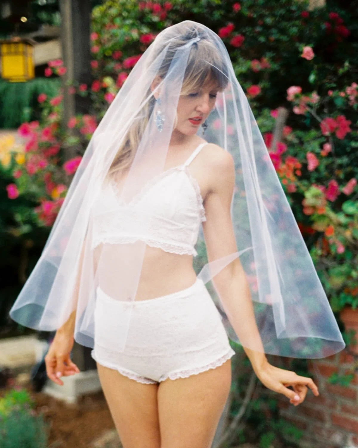 A woman in white vintage wedding lingerie and a veil standing outdoors in front of pink flowers.