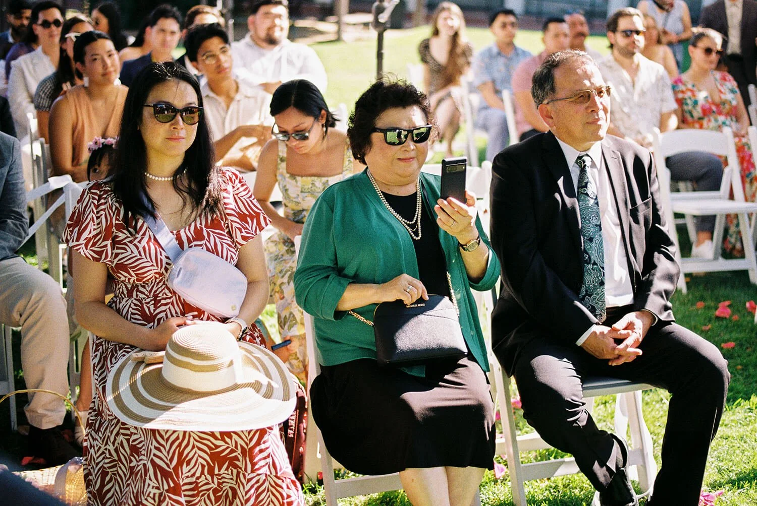 Group of people seated outdoors at a garden wedding, including women wearing sunglasses and a man in a suit, some are using their phones to take photos, with a background of more attendees and a grassy area.