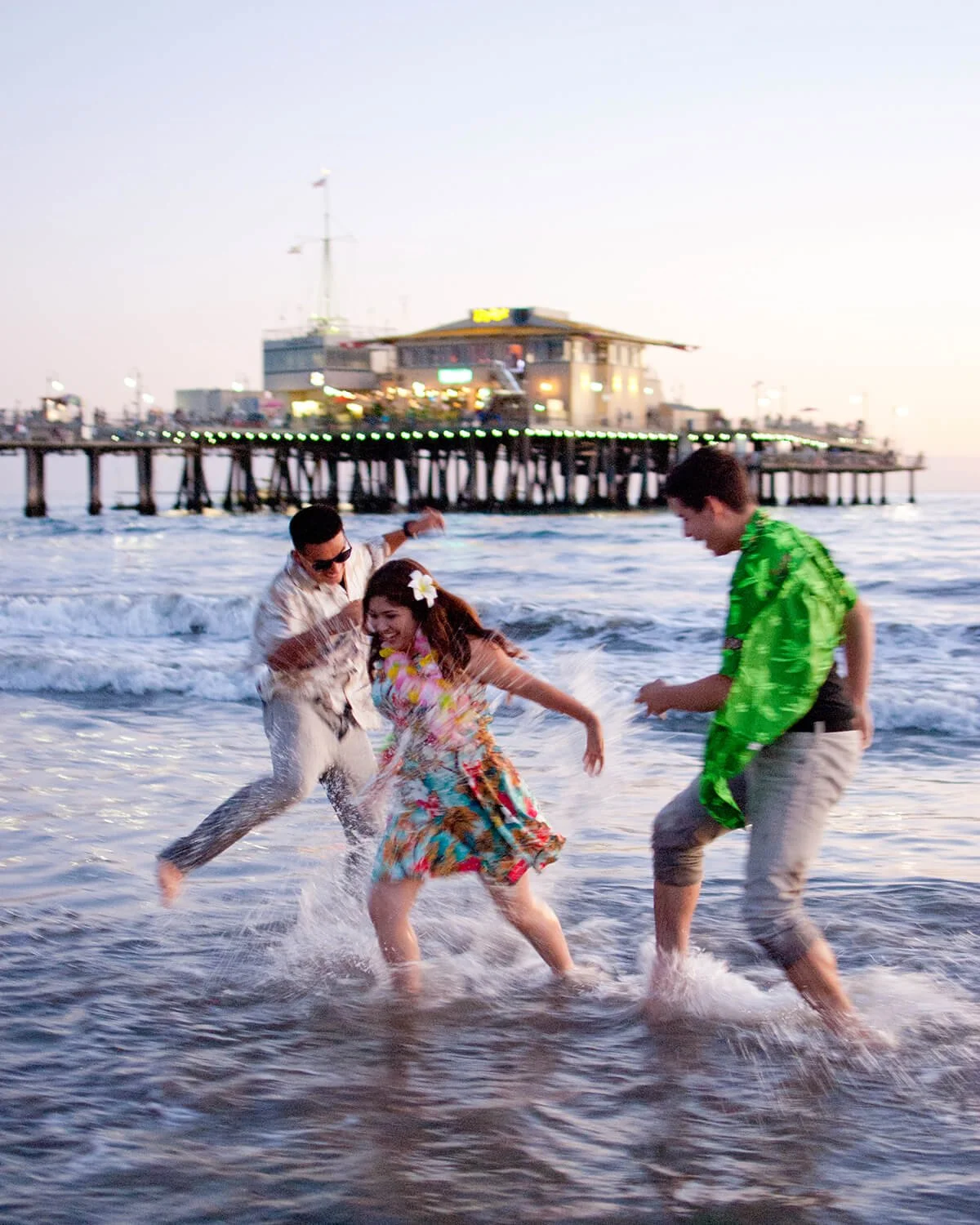 Three people playing and splashing in the ocean near a pier with a building in the background during sunset.