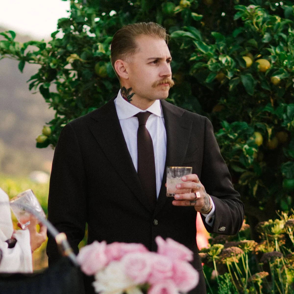 A man dressed in a black suit with a white shirt and black tie, holding a glass, standing outdoors near a lush green orchard or garden.