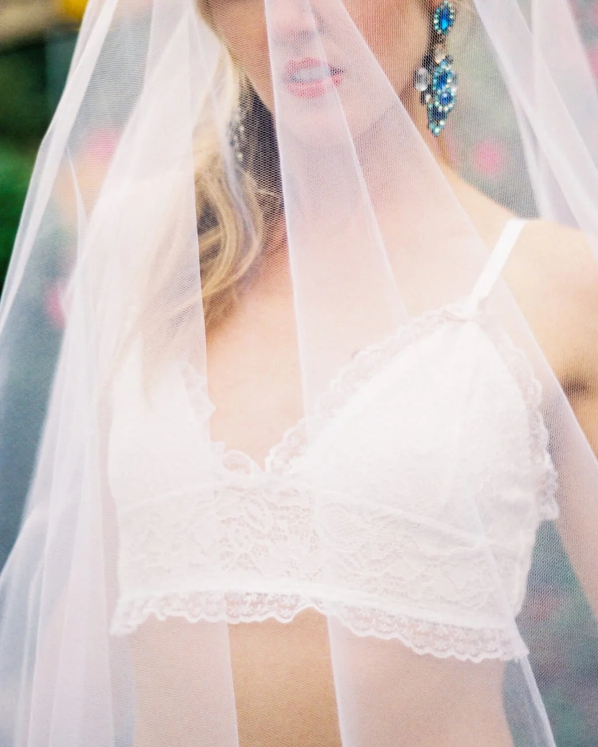 Close-up of a woman wearing a white lace bra and a veil, with large blue earrings, partially obscured by the veil.