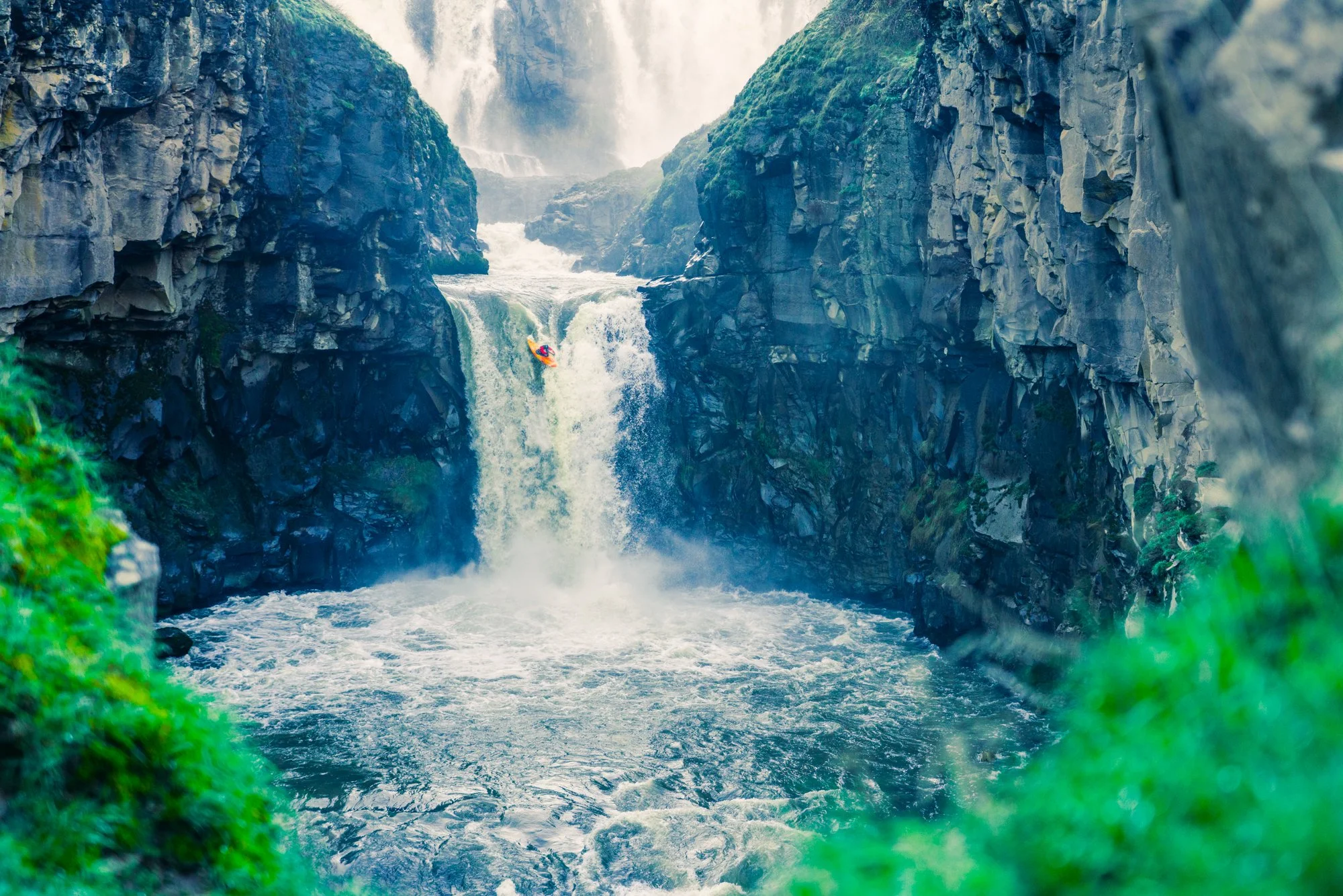 Atmospheric misty outdoor sports photography of a kayaker dropping a large waterfall. The image is saturated and blue, with grass in the foreground. This photo was featured in an Outside Magazine article about capturing the perfect action adventure s