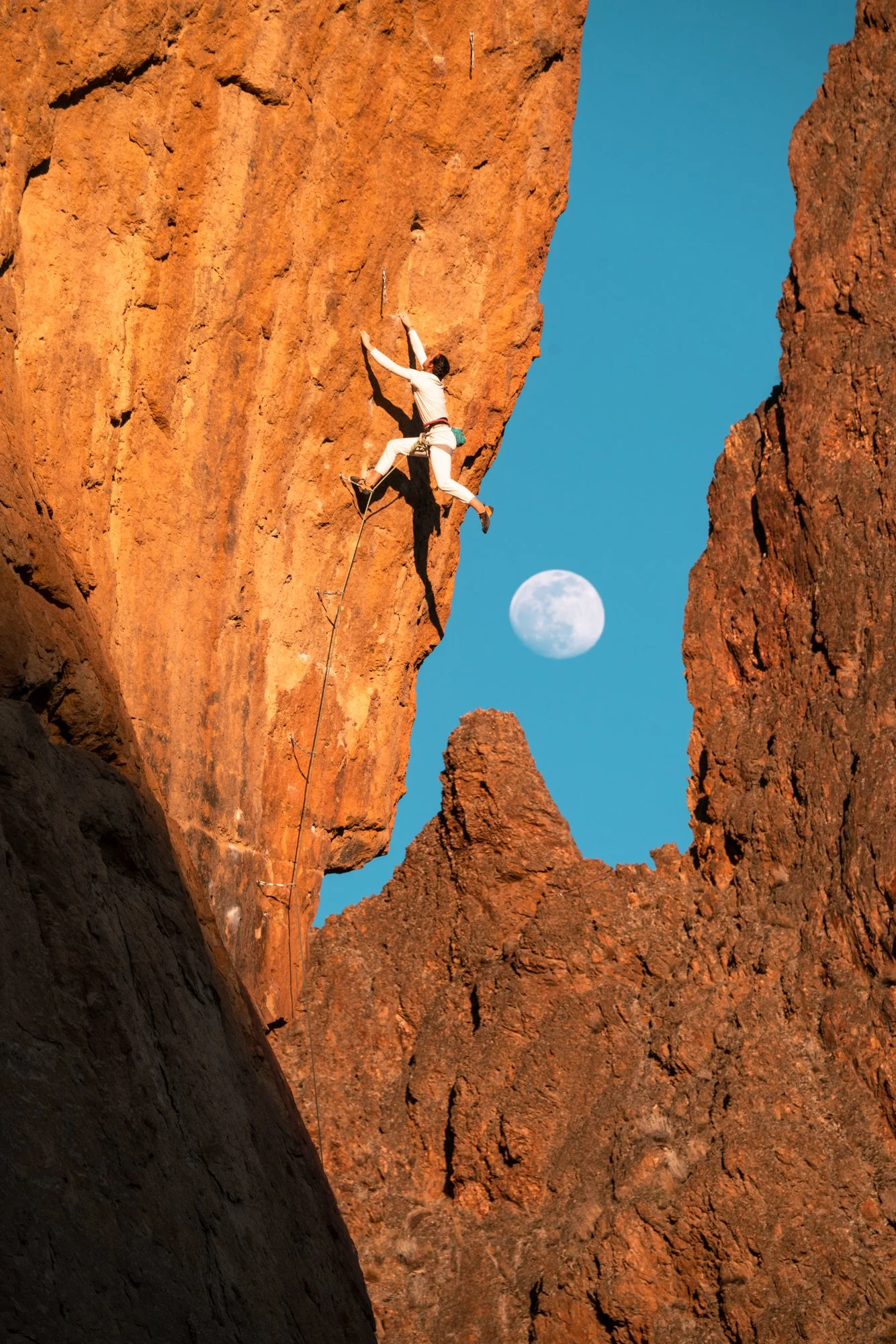 Rock climber scaling a steep rock face during golden hour with a clear blue sky as the full moon rises over the horizon. This photo was taken on Time to Power 5.12c at Smith Rock State Park in Central Oregon.