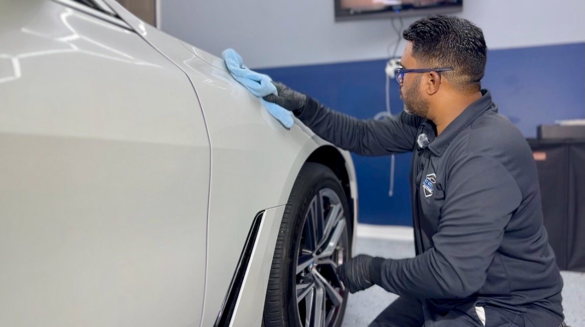 A man wearing glasses, black gloves, and a gray uniform is polishing a white car with a blue cloth in an auto detail shop.