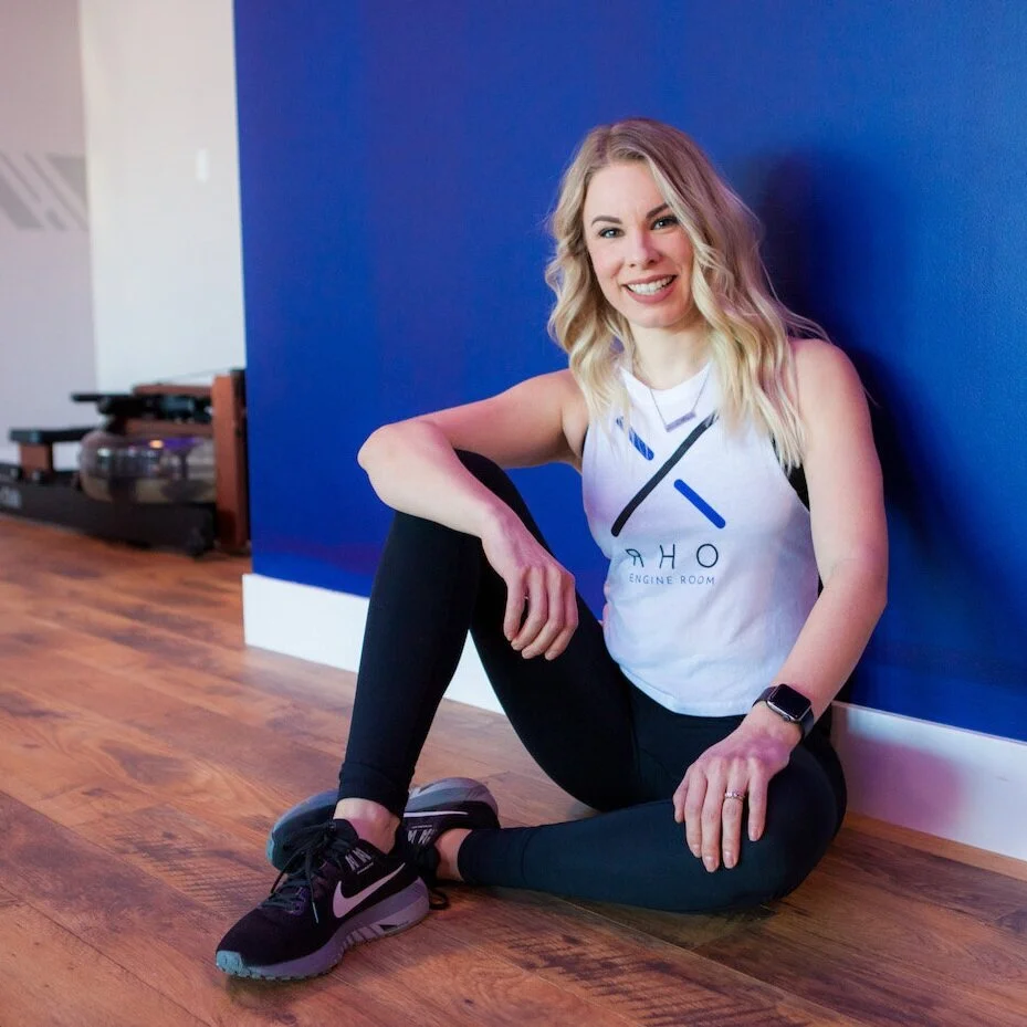 Coach Shawna sitting in front of a blue wall with a Rho shirt on, with a rower in the background