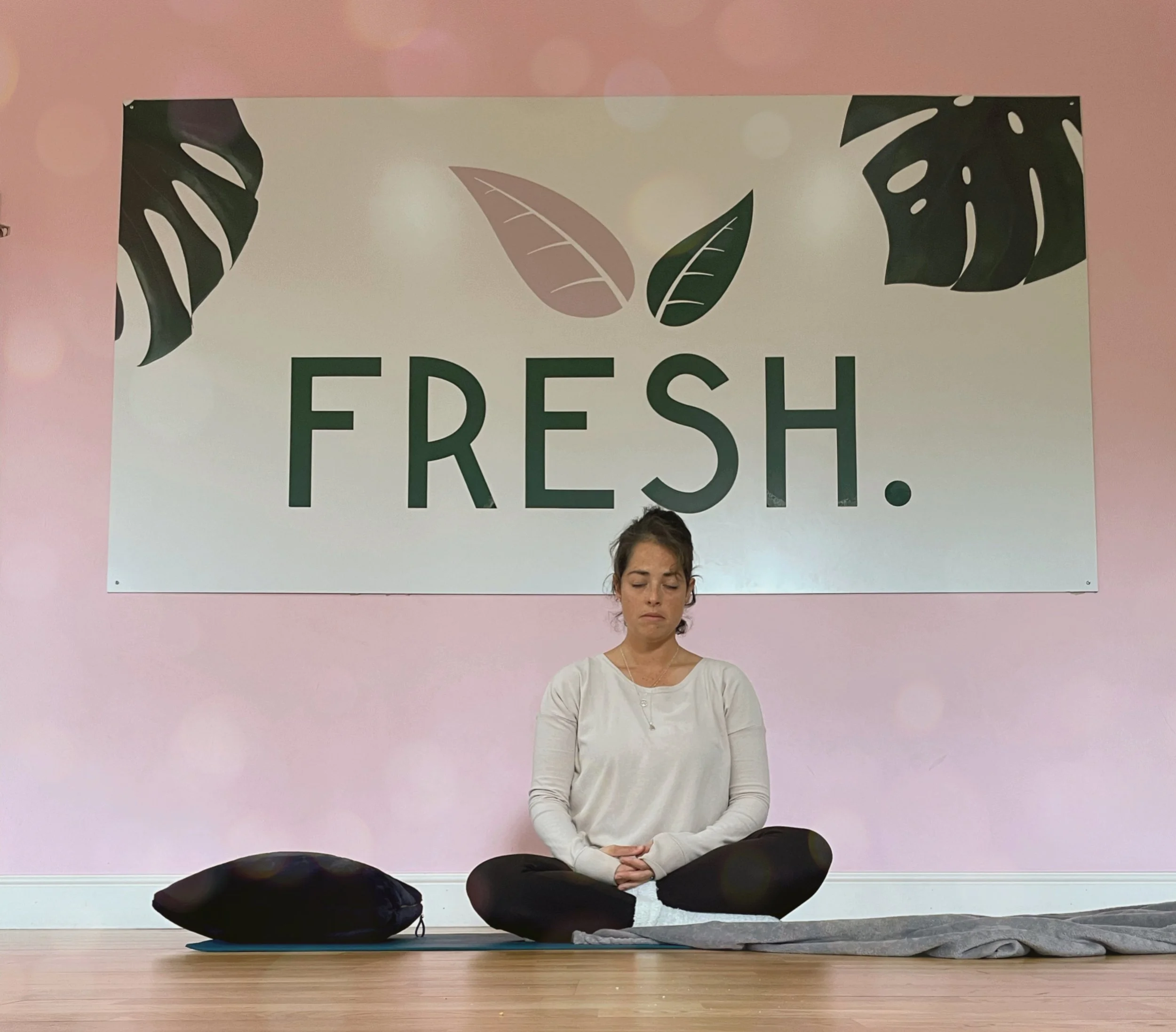A woman doing a meditation or yoga practice while sitting cross-legged on a mat in a room with a pink wall. There is a large sign on the wall with the word 'FRESH.' and some leaves.