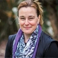 Close-up of Johanna Hooper,  woman with brown hair, wearing a dark jacket and a purple scarf, smiling outdoors.
