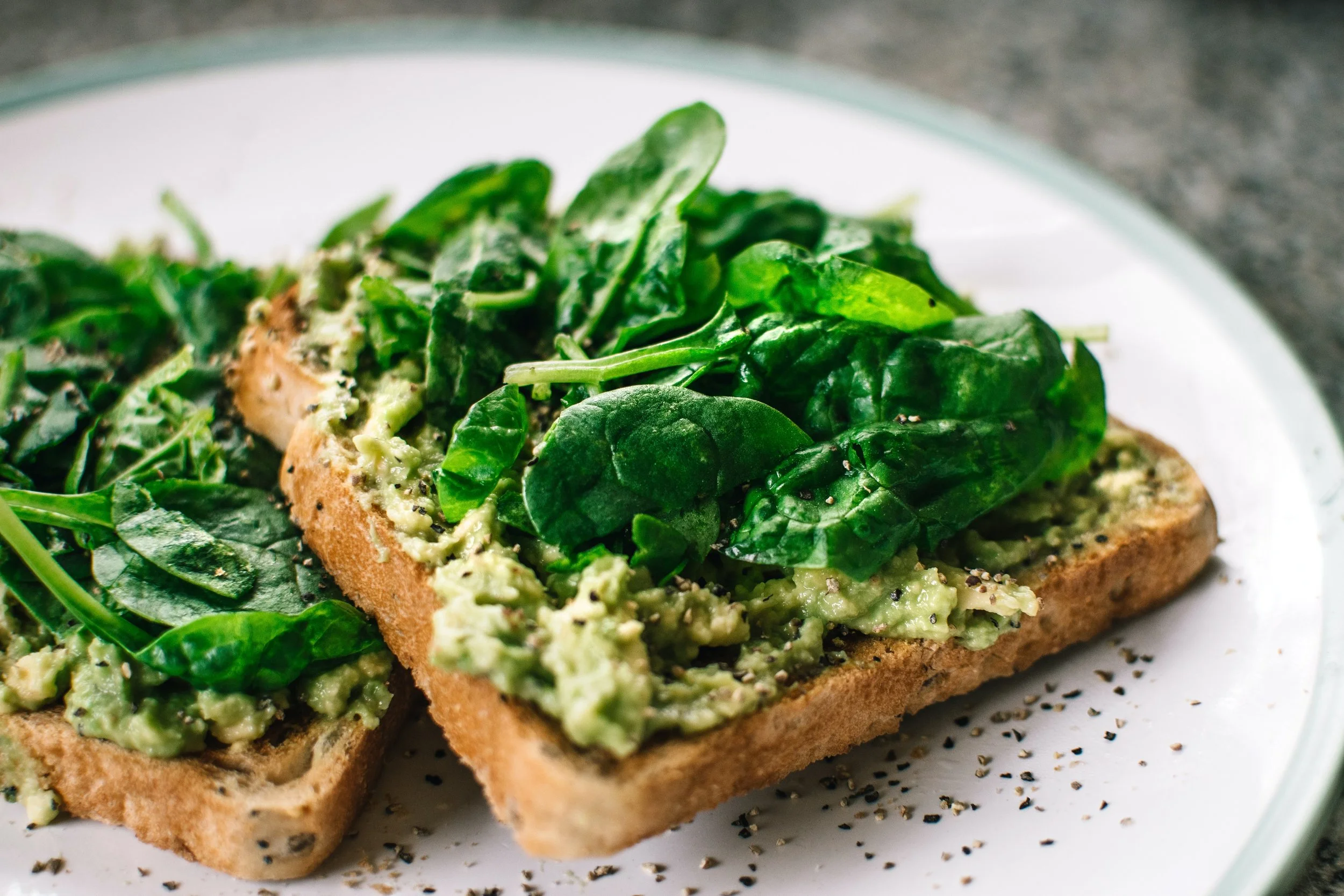 Avocado toast topped with fresh spinach leaves on a white plate.