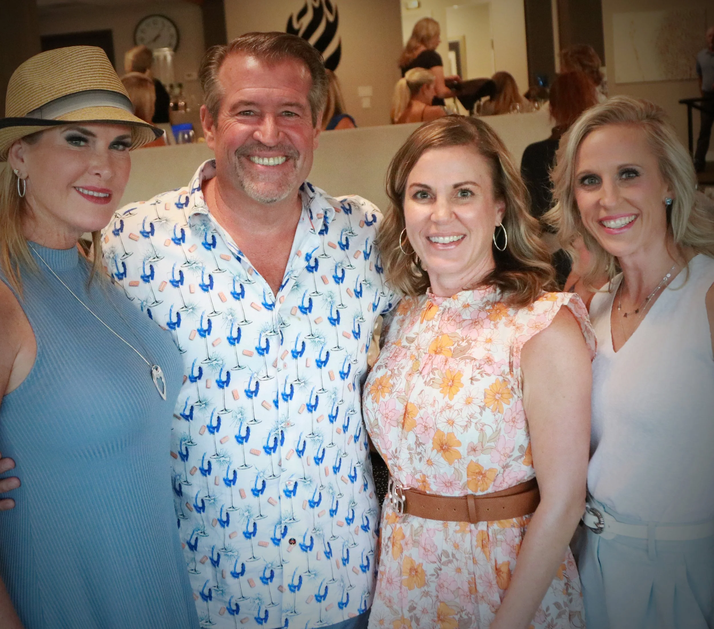 Four people, three women and one man, smiling at a social gathering, with a bar and other people in the background.