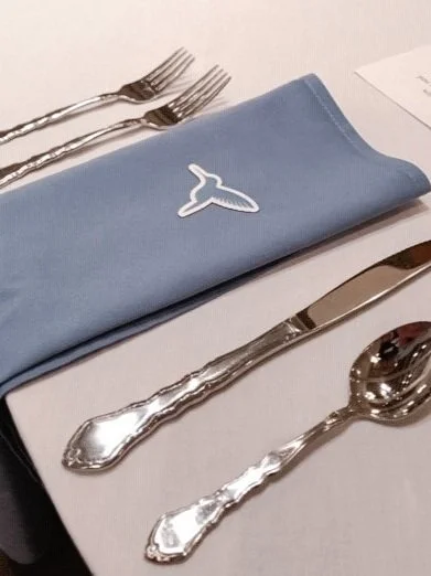 Place setting with forks, knives, and spoons on a white table, a blue napkin with a whale logo.