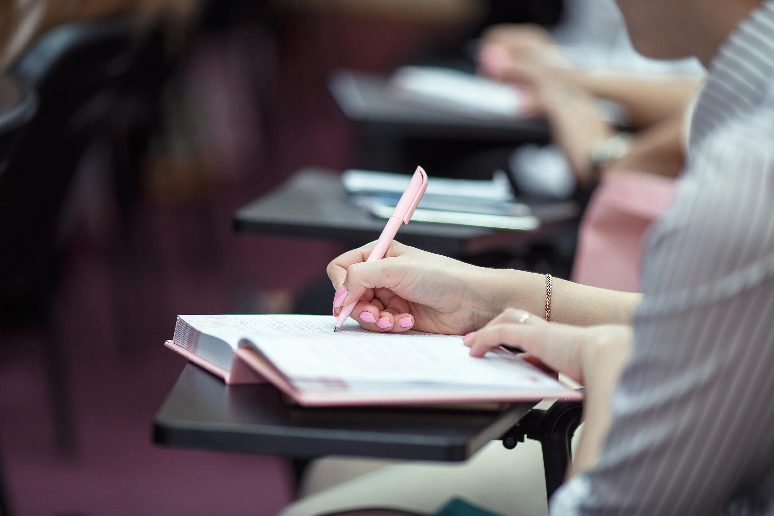 A person in a striped shirt writing in a pink notebook with a pink pen at a desk, with other people and notebooks visible in the background.