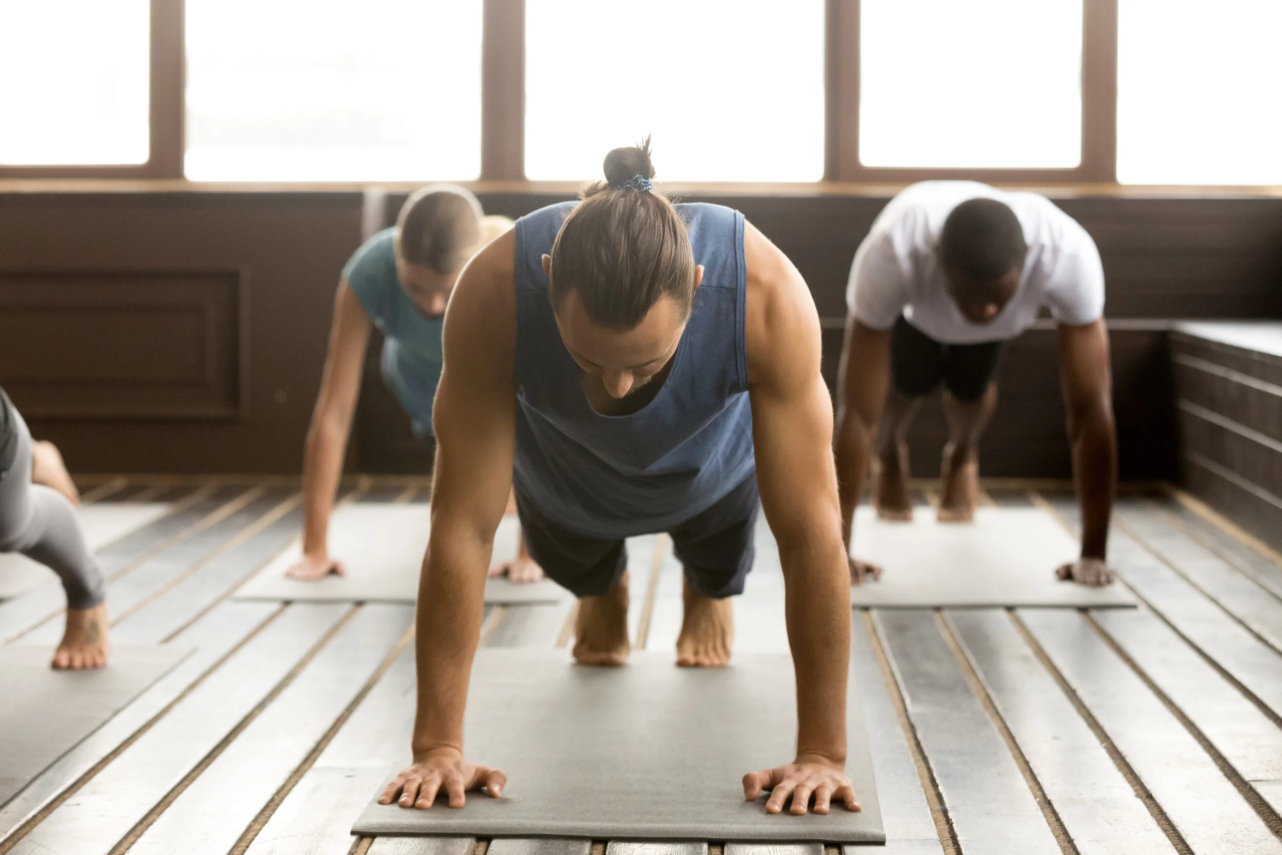 People doing a yoga plank pose on mats in a room with large windows.