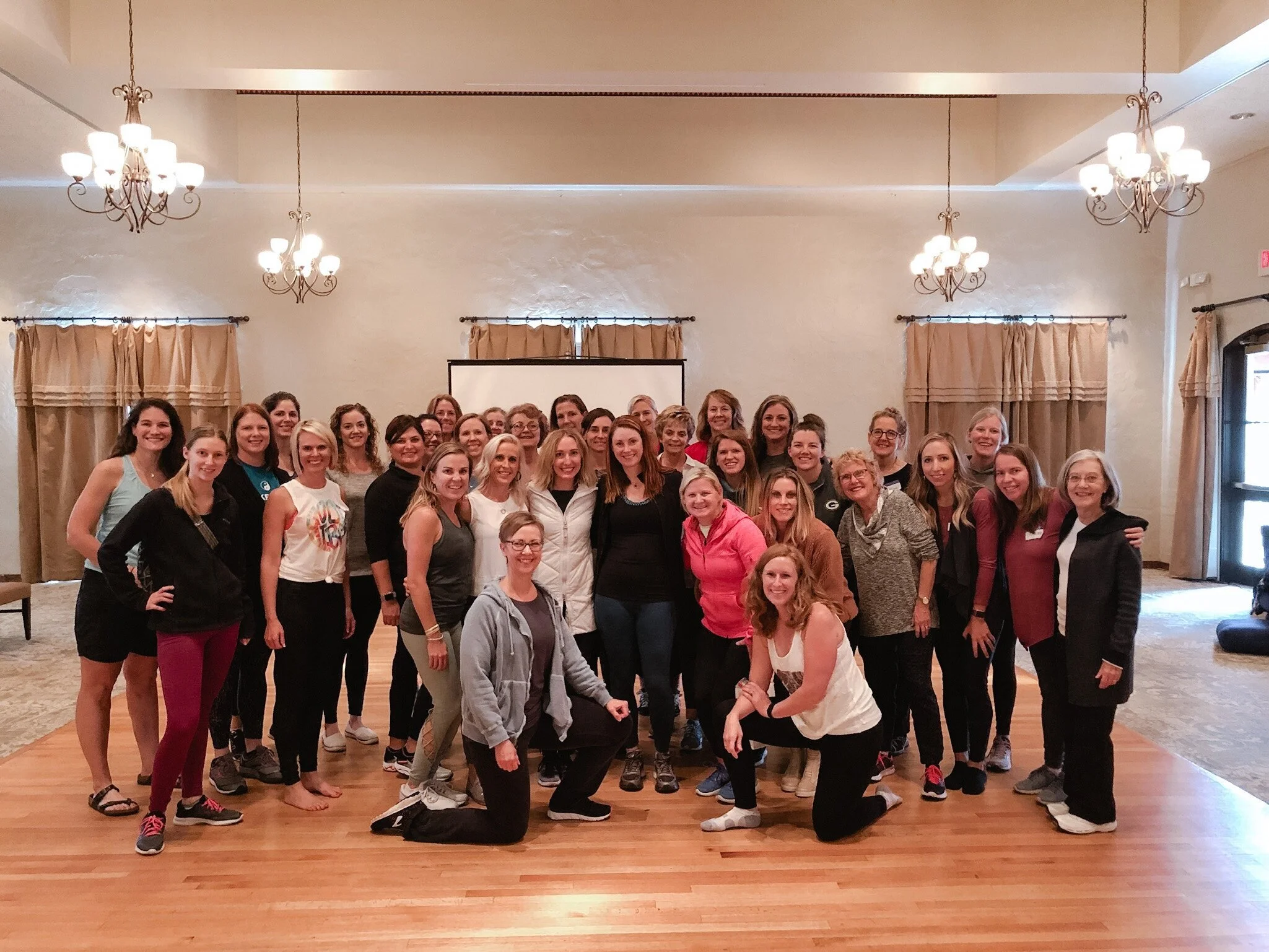 A large group of women gathered together indoors, posing for a group photo in a room with chandeliers, beige curtains, and wood flooring.