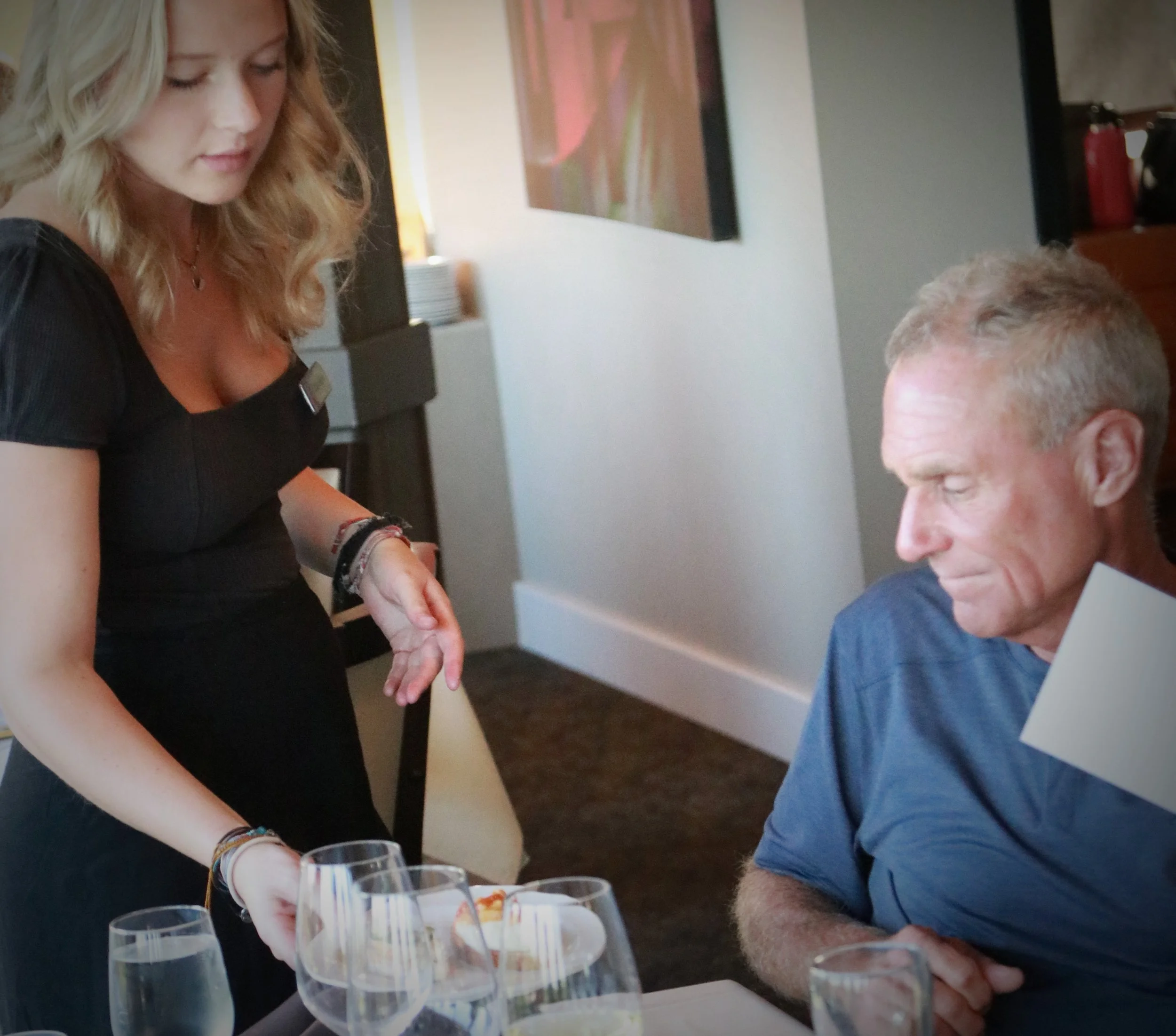 A woman wearing a black dress is serving food to an older man with short gray hair, seated at a table with multiple wine glasses in a restaurant or dining setting.