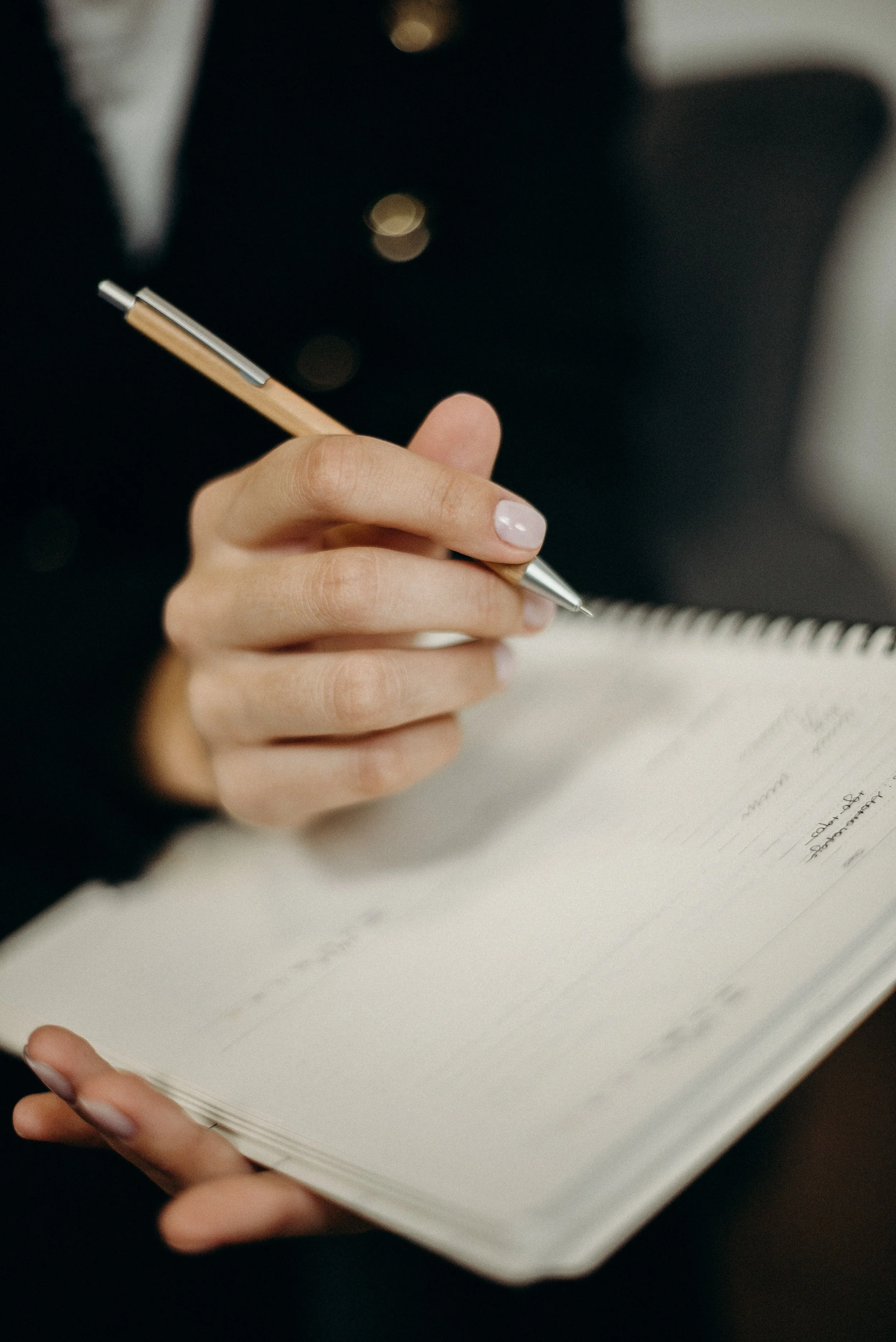 A woman in black clothing holding a pen and writing in a spiral notebook.