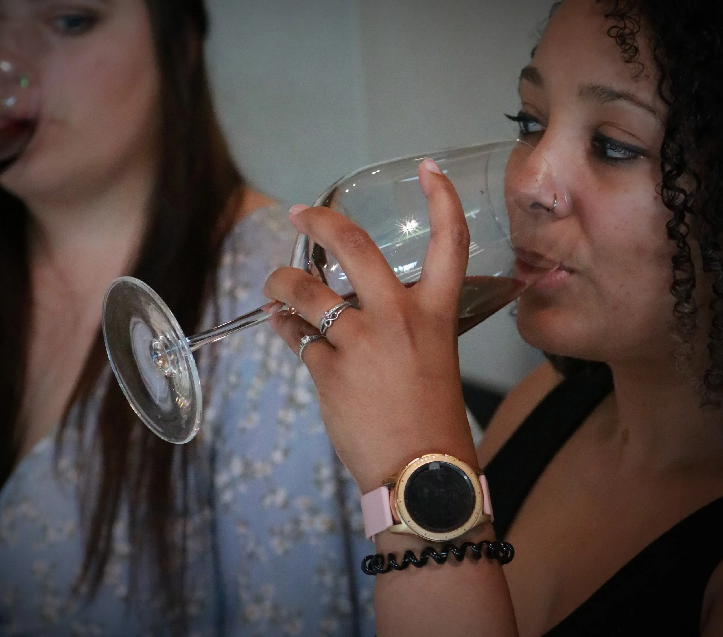 A woman with curly hair and a nose ring drinking red wine from a wine glass during a social gathering.