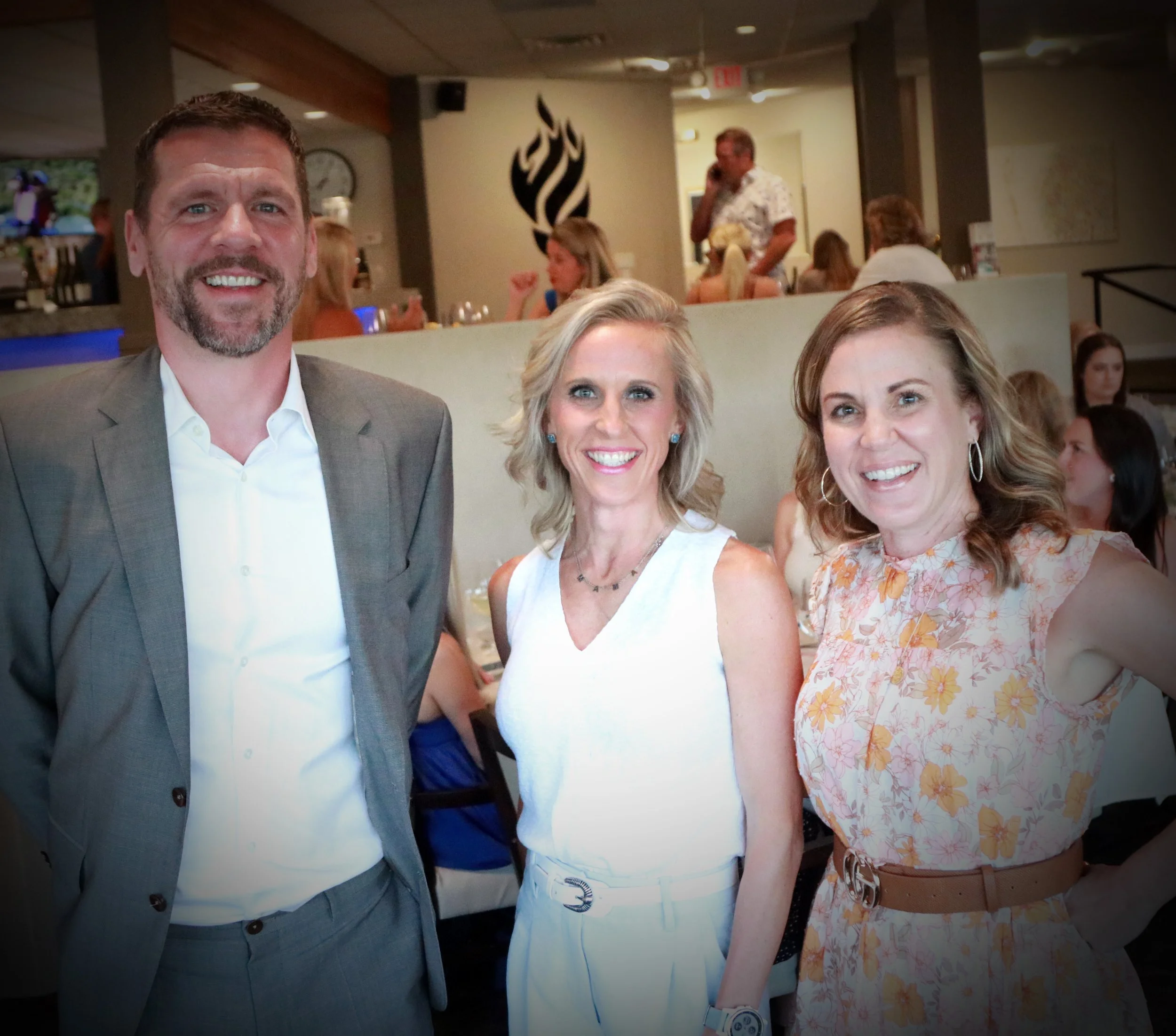 Three smiling adults, two women and one man, standing together indoors at a social event; the man is wearing a gray suit, the woman in the middle wears a white sleeveless top, and the woman on the right wears a floral dress.