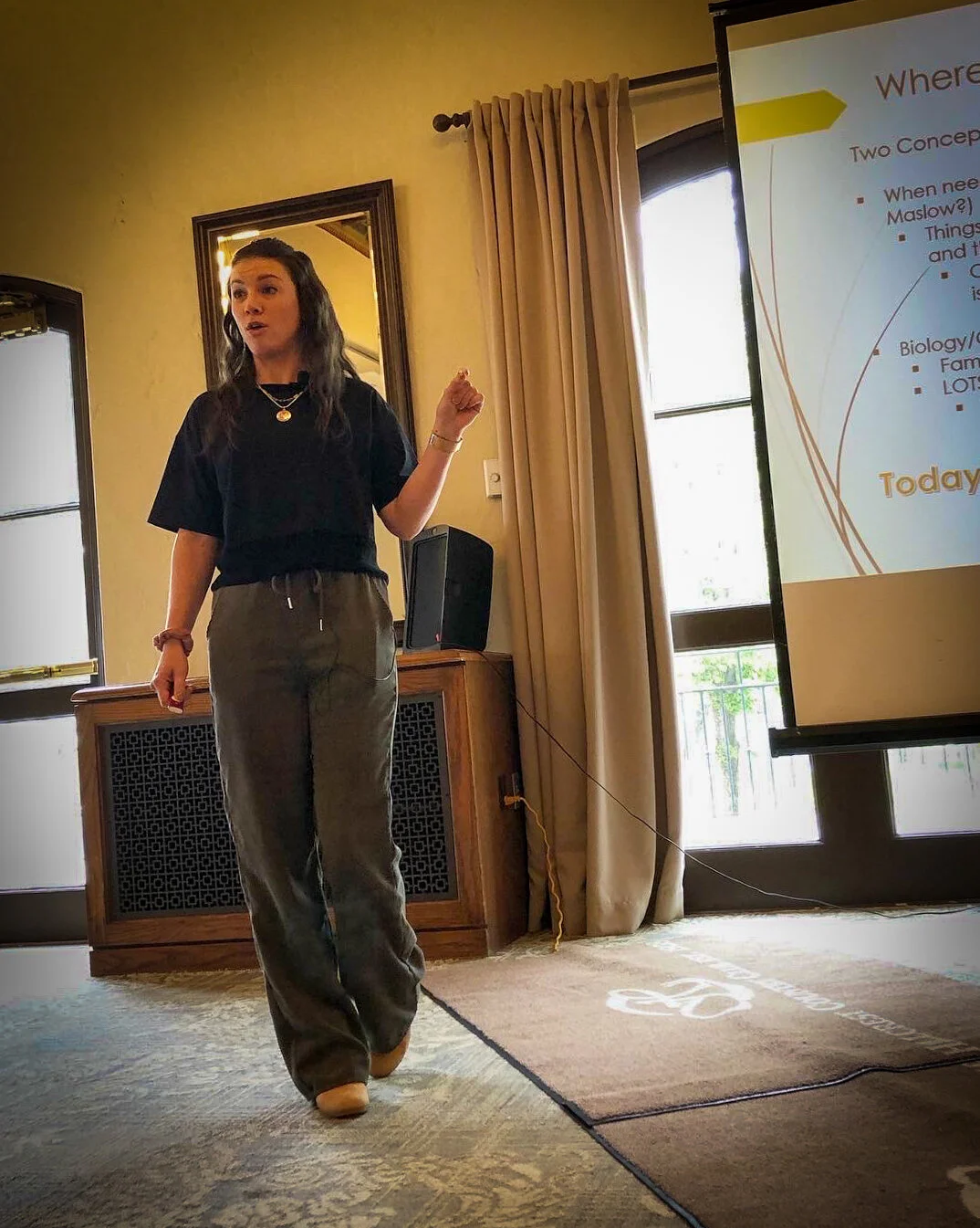 A woman giving a presentation next to a screen with slides, standing in a room with beige walls, curtains, and windows, pointing towards the slides.