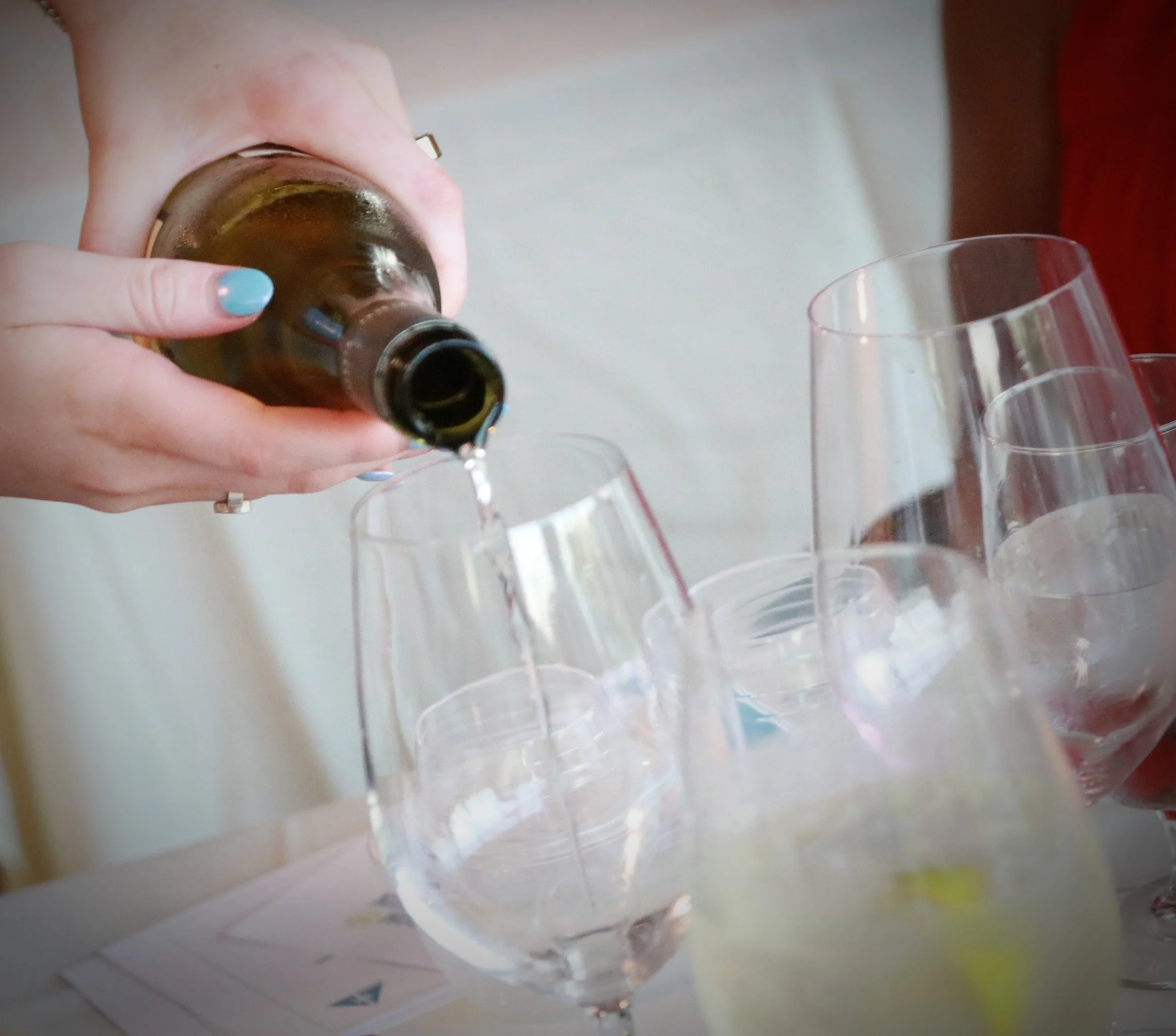 Person pouring a drink from a brown bottle into a wine glass, with several other glasses on the table.