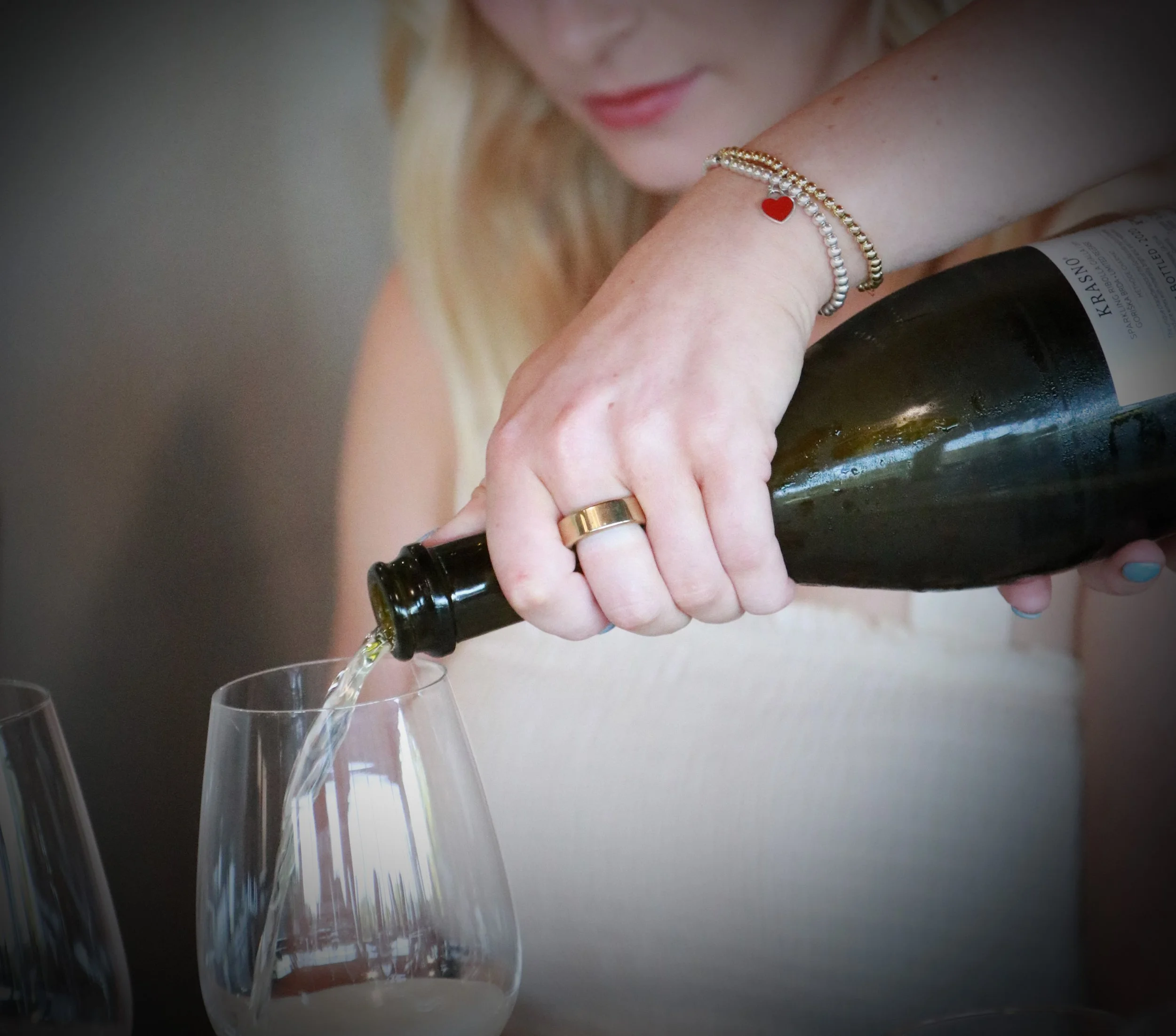 A woman pours white wine from a bottle into a wine glass, wearing a gold wedding ring and a jewelry bracelet.