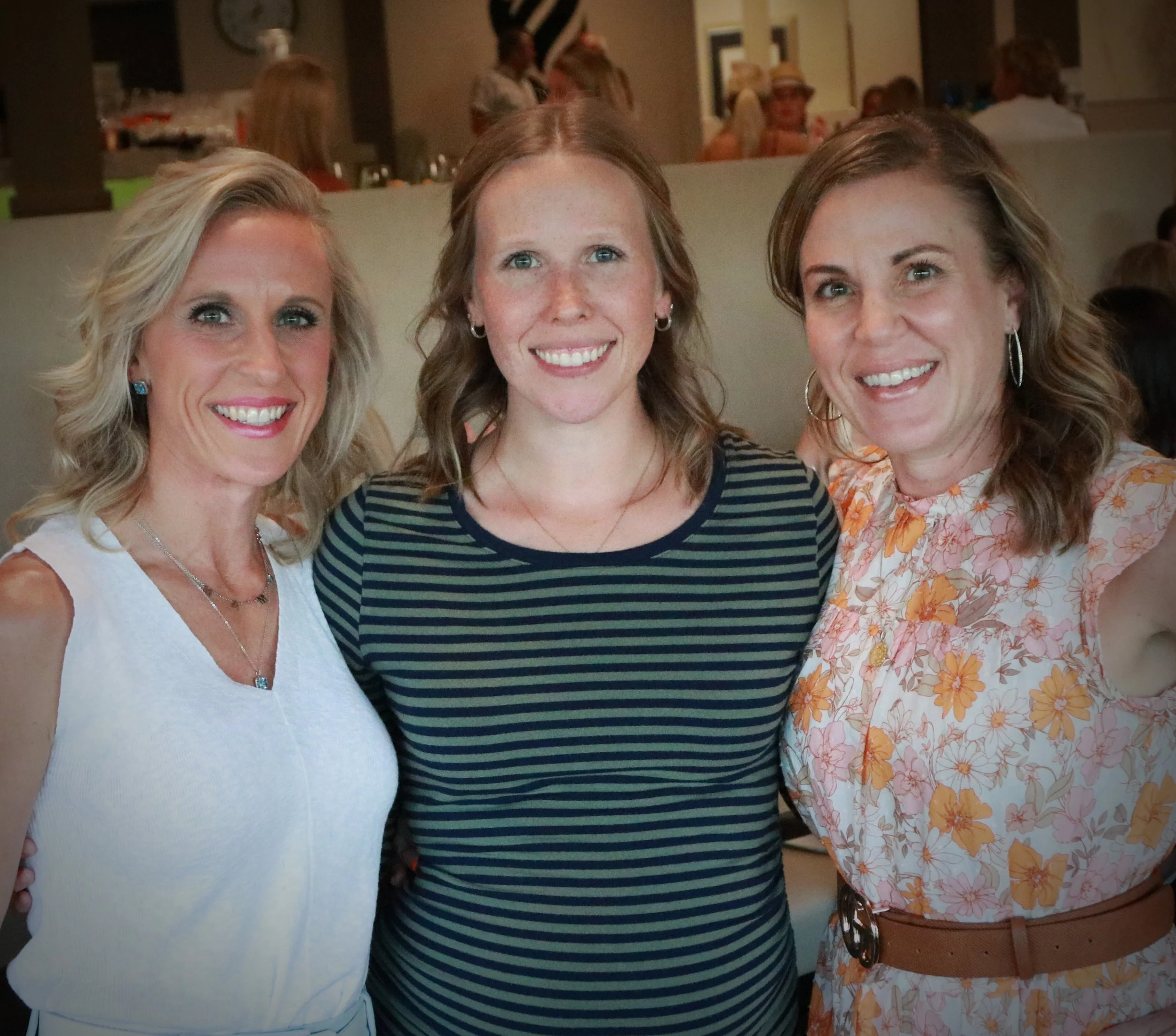Three women standing close together, smiling at the camera, in a restaurant or cafe setting with other people in the background.