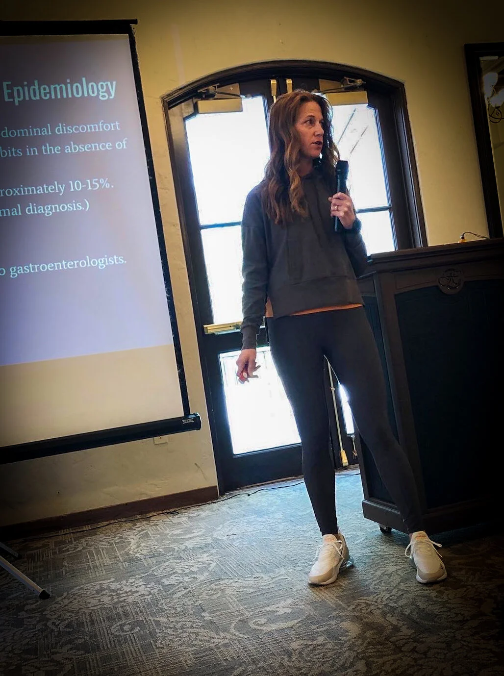 A woman giving a presentation in a conference room, holding a microphone, with a slide titled 'Epidemiology' projected on a screen.