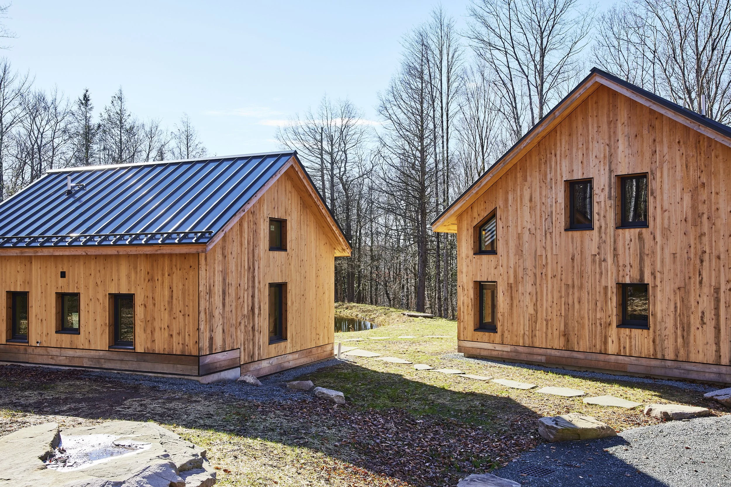 Two wooden houses with sloped roofs are situated outdoors, with a small pond and leafless trees in the background, under a clear blue sky.