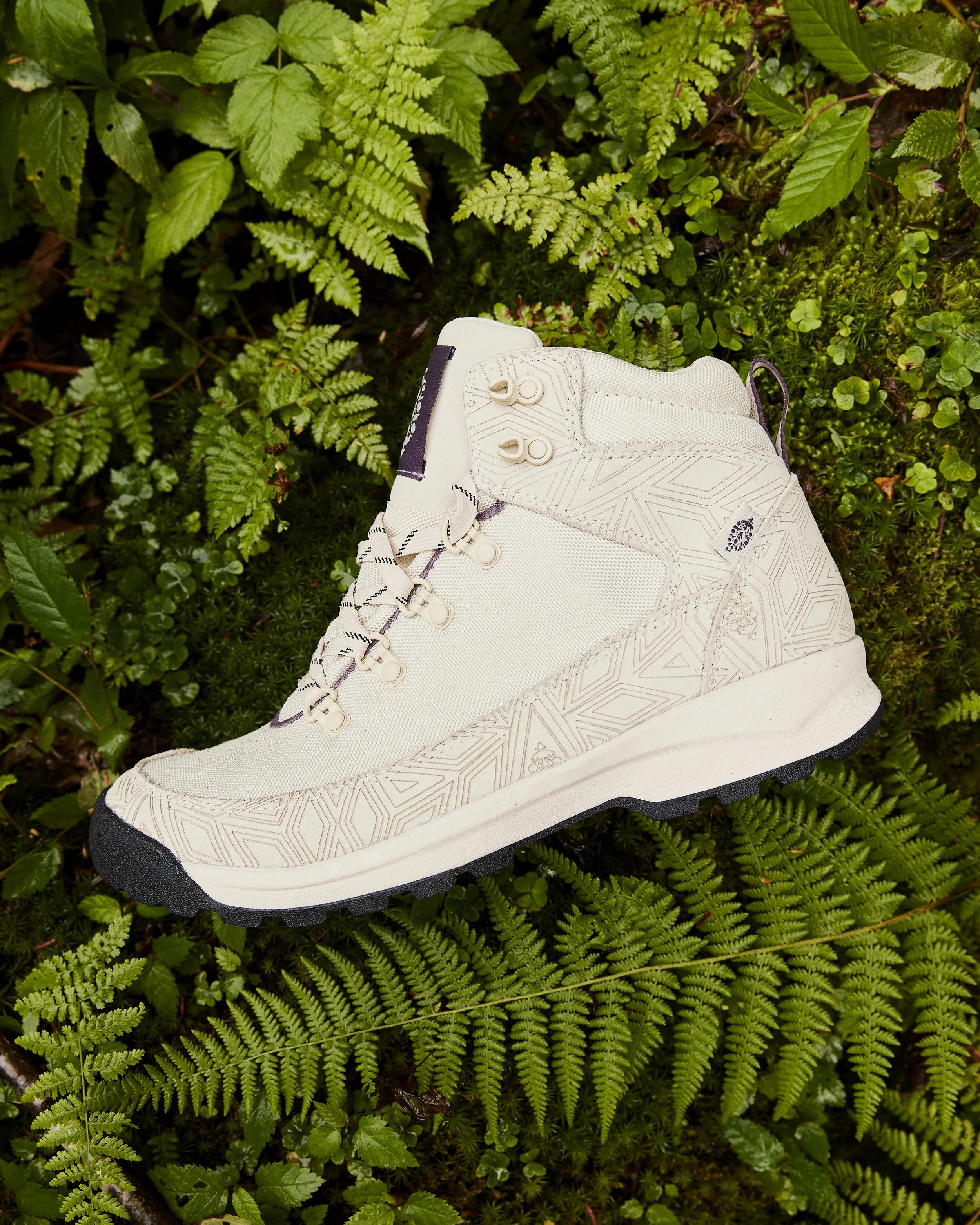 A white hiking shoe with patterned design, laces, and a rugged sole, resting on green ferns and foliage.