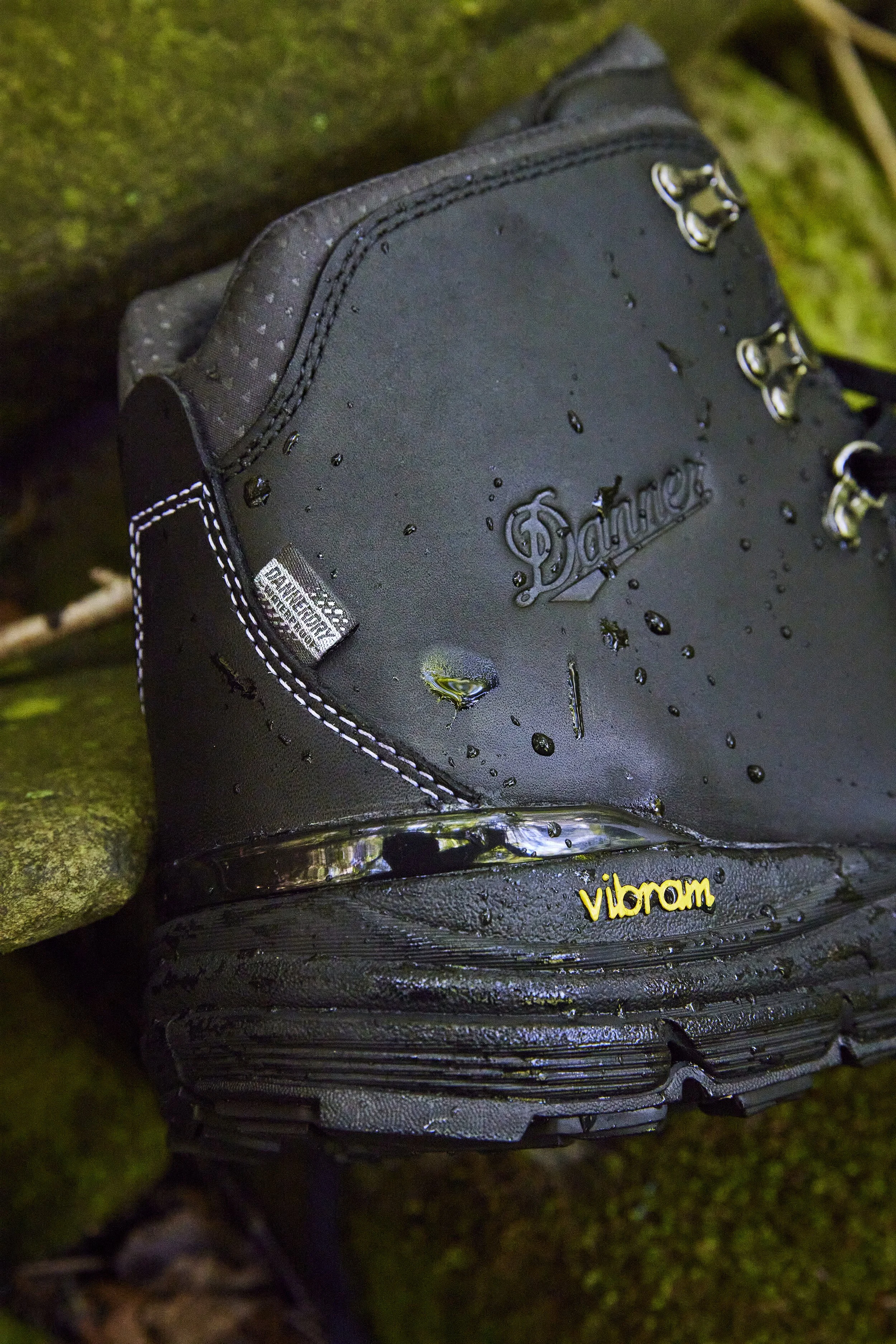 A black hiking boot with water droplets on it, resting on a mossy rock. The boot has a 'Vibram' logo near the sole and a 'Danner' logo on the side.