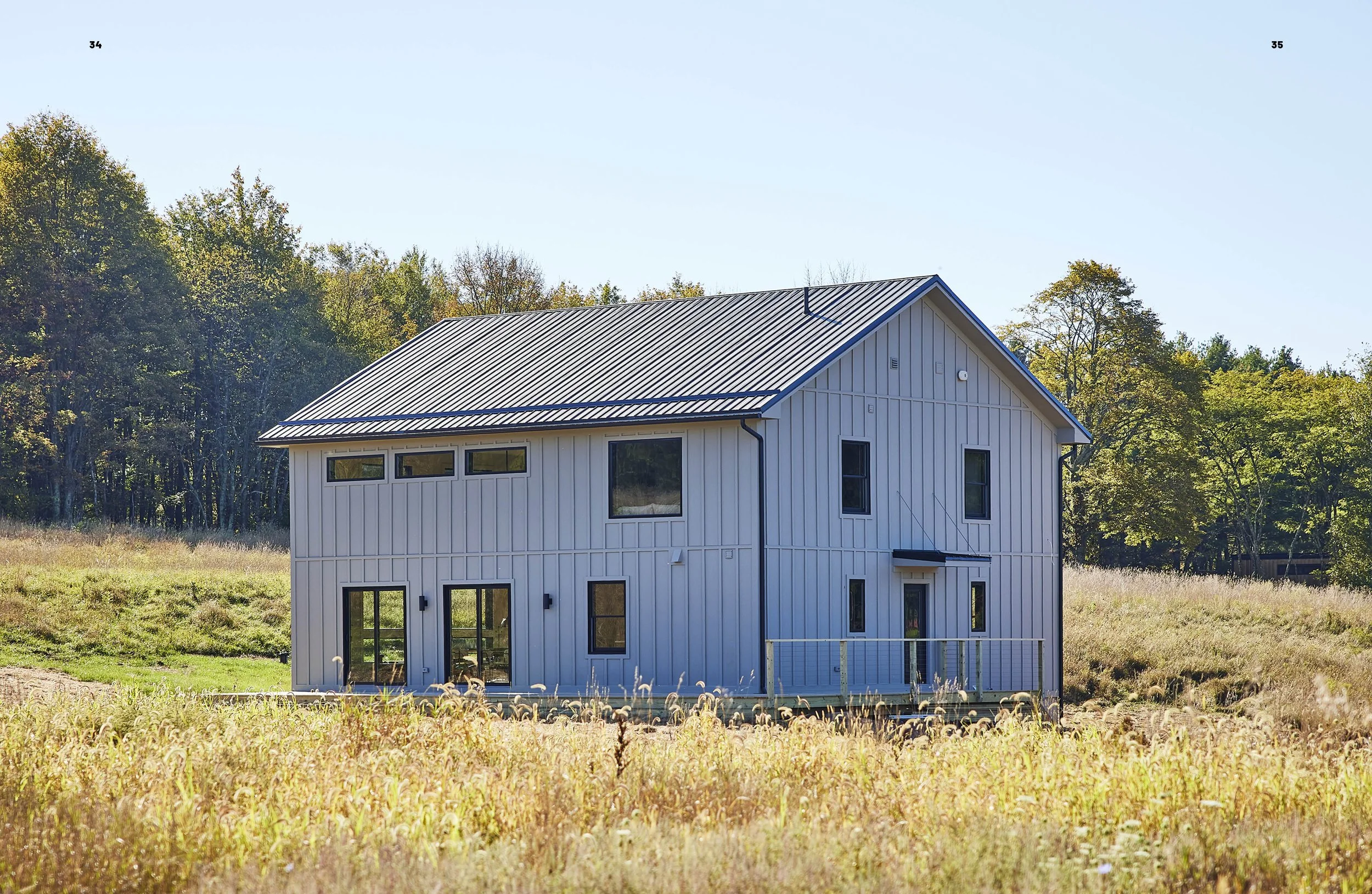 A two-story modern house with white vertical siding, black window frames, and a metal roof, situated on a grassy field with trees in the background.