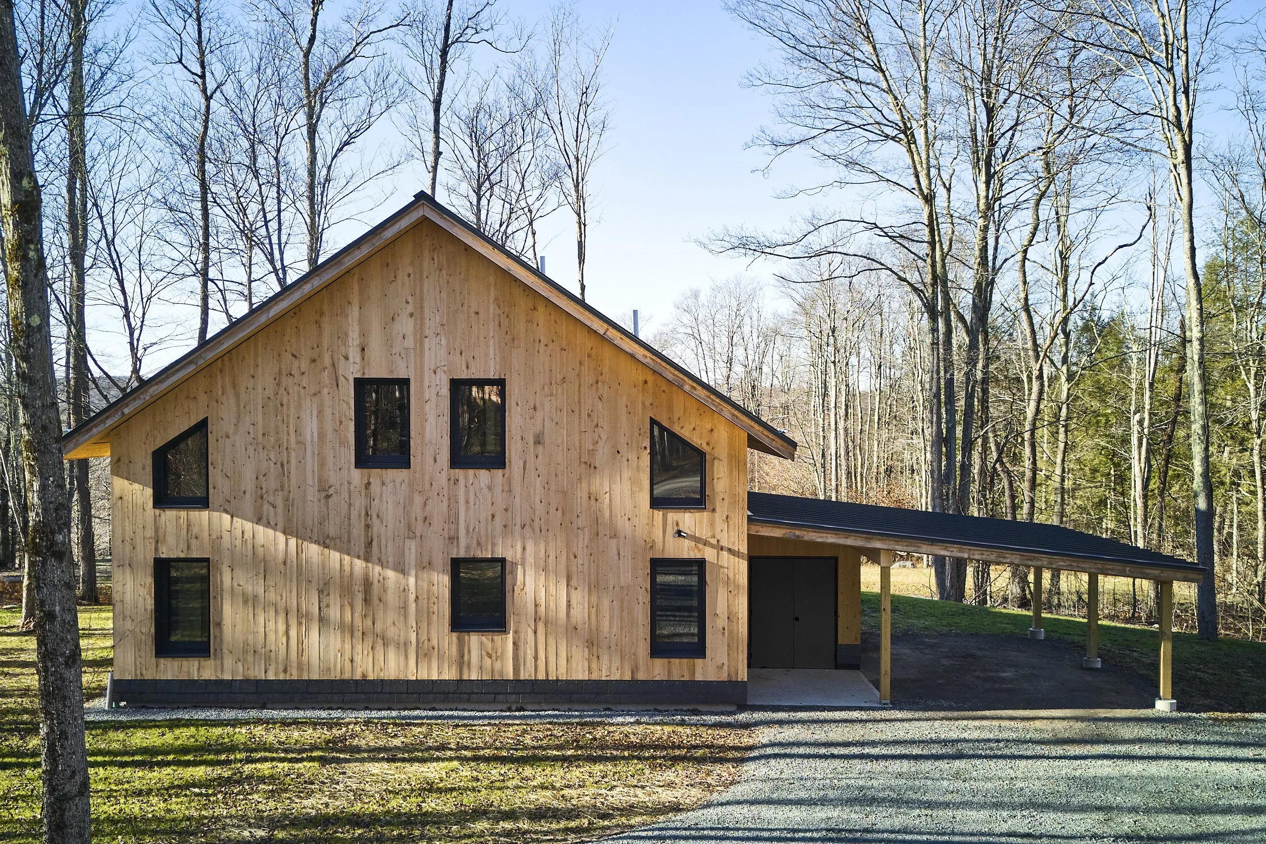 A modern wooden house with multiple rectangular and angled windows, situated in a wooded area with leafless trees in the background and a gravel driveway in front.