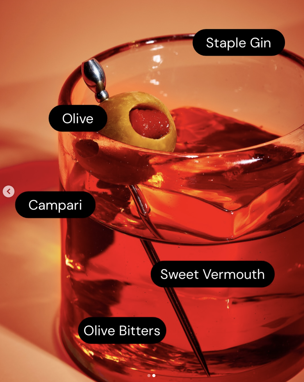 Close-up of a cocktail in a glass with a green olive with a red pimento, an olive pick, and a large ice cube. The drink contains sweet vermouth, campari, olive bitters, and is labeled as a staple gin cocktail.