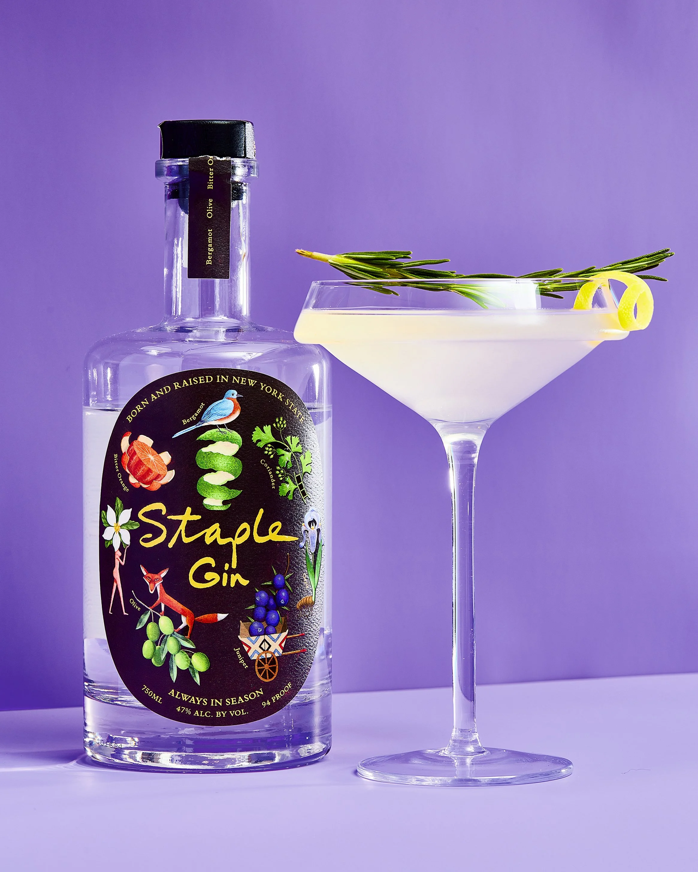A bottle of Staple Gin beside a cocktail glass garnished with a sprig of rosemary and lemon peels, set on a purple background.
