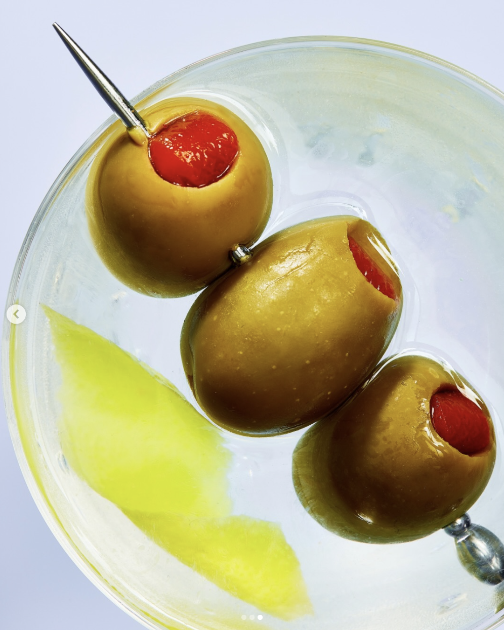 Three green olives stuffed with red pimiento, skewered on cocktail picks, floating in a martini glass with an olive brine and a lemon wedge.
