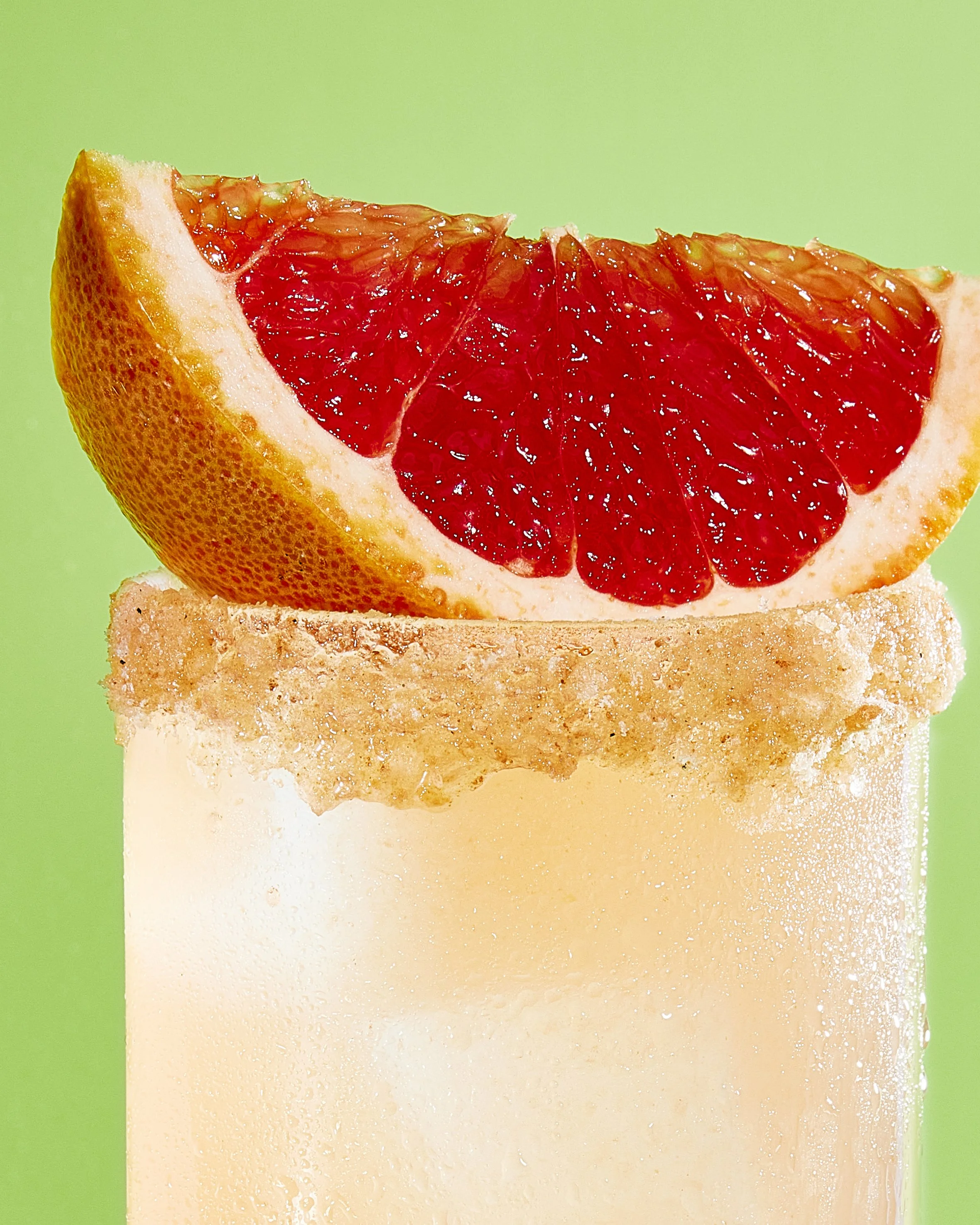 Close-up of a grapefruit slice on top of a creamy drink in a glass with a salted rim, against a green background.