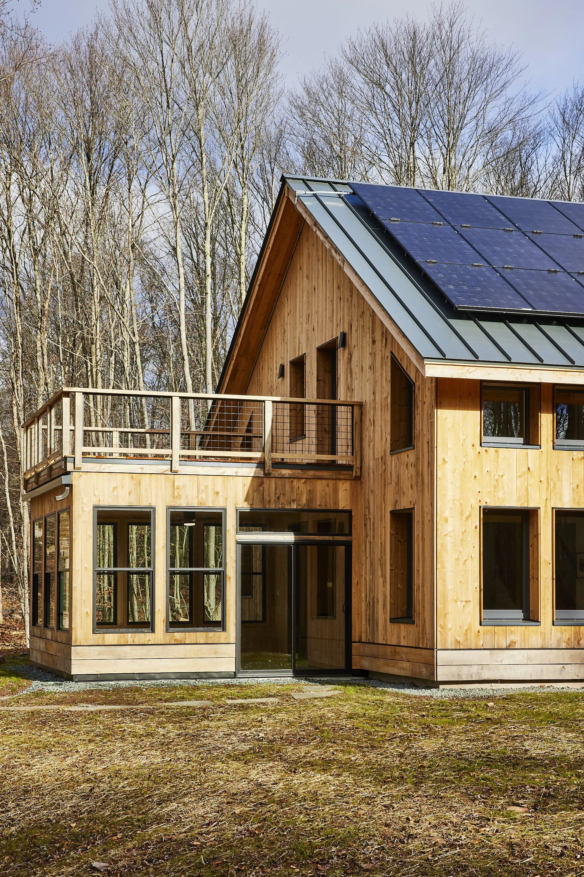A modern wooden house with large windows, a glass door, and solar panels on the roof, surrounded by bare trees and a grassy yard.