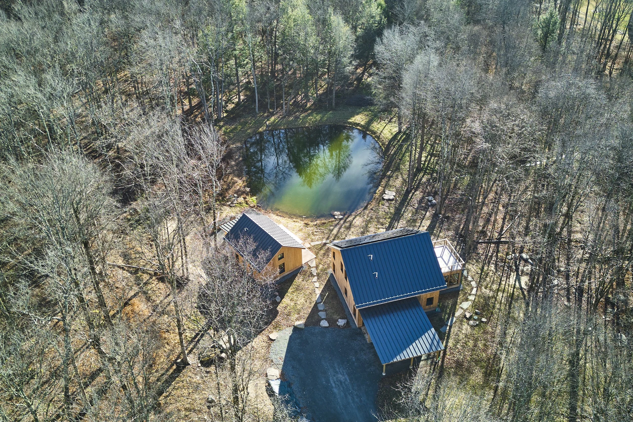 Aerial view of two small cabins with dark metal roofs surrounded by trees, near a small pond in a wooded area, in early spring.