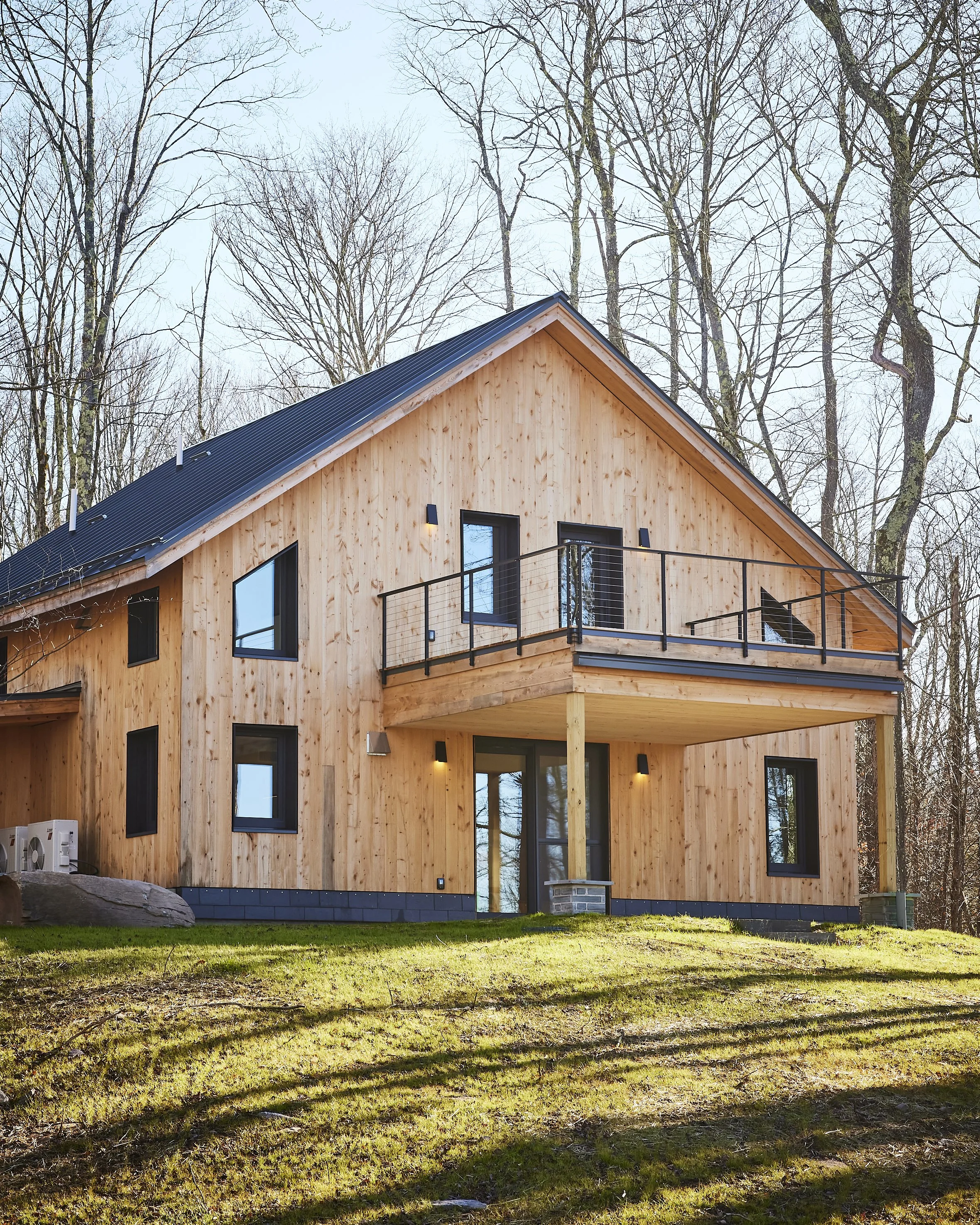 A two-story wooden house with a black metal roof and a balcony, situated in a wooded area with leafless trees in the background and a grassy lawn in the foreground.