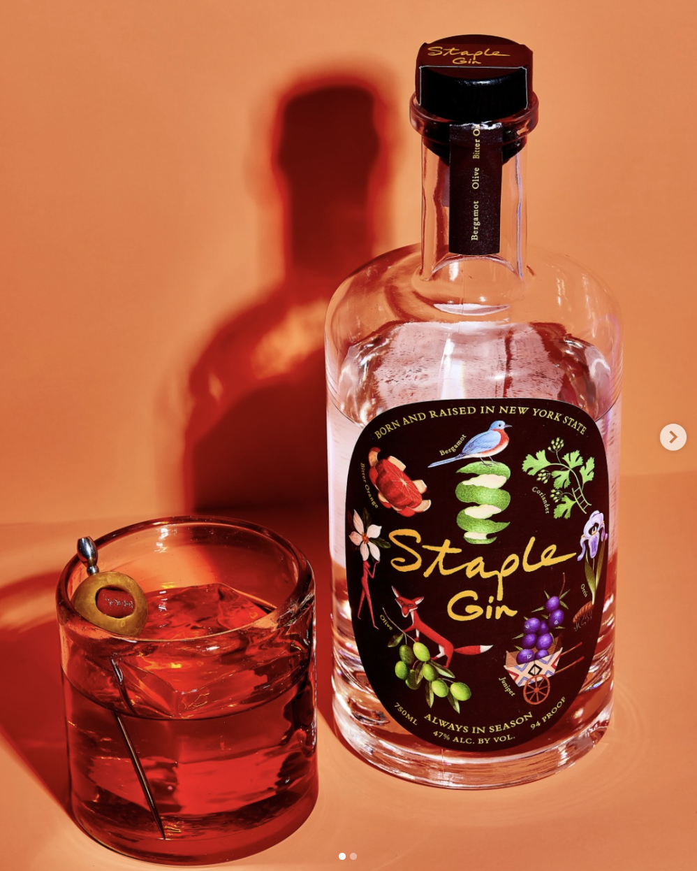 A bottle of Grown and Raised in New York State Staple Gin with a glass of red-colored cocktail garnished with an olive on a cocktail pick, set against an orange background with a shadow of the bottle.