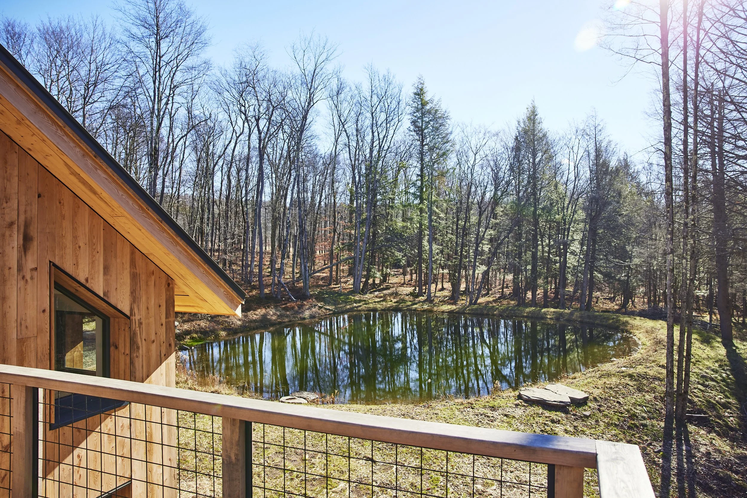 A wooden house with a deck, overlooking a small pond surrounded by trees and woods on a sunny day.