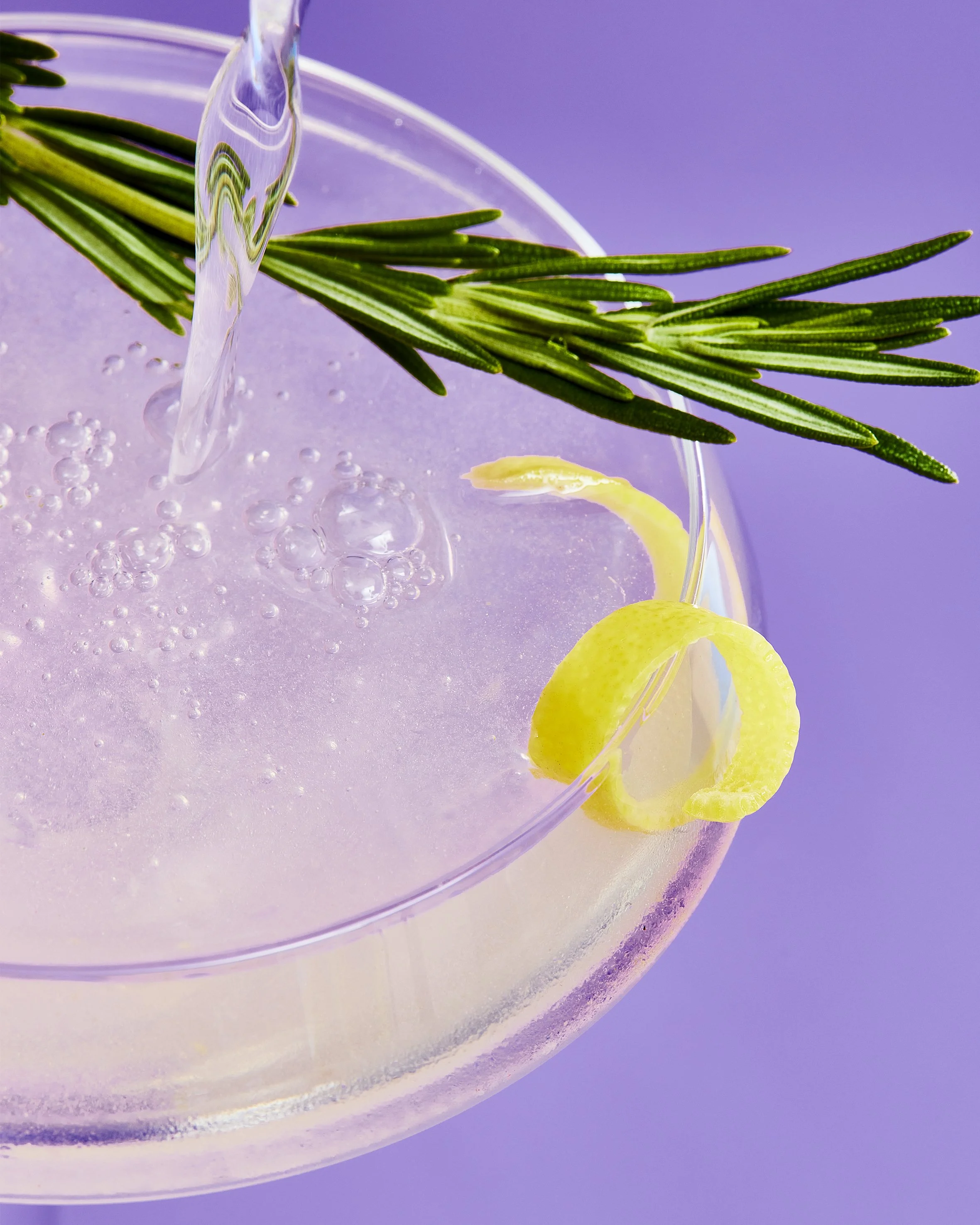 Close-up of a cocktail glass with a lemon peel and rosemary garnish, bubbly liquid, and a purple background.
