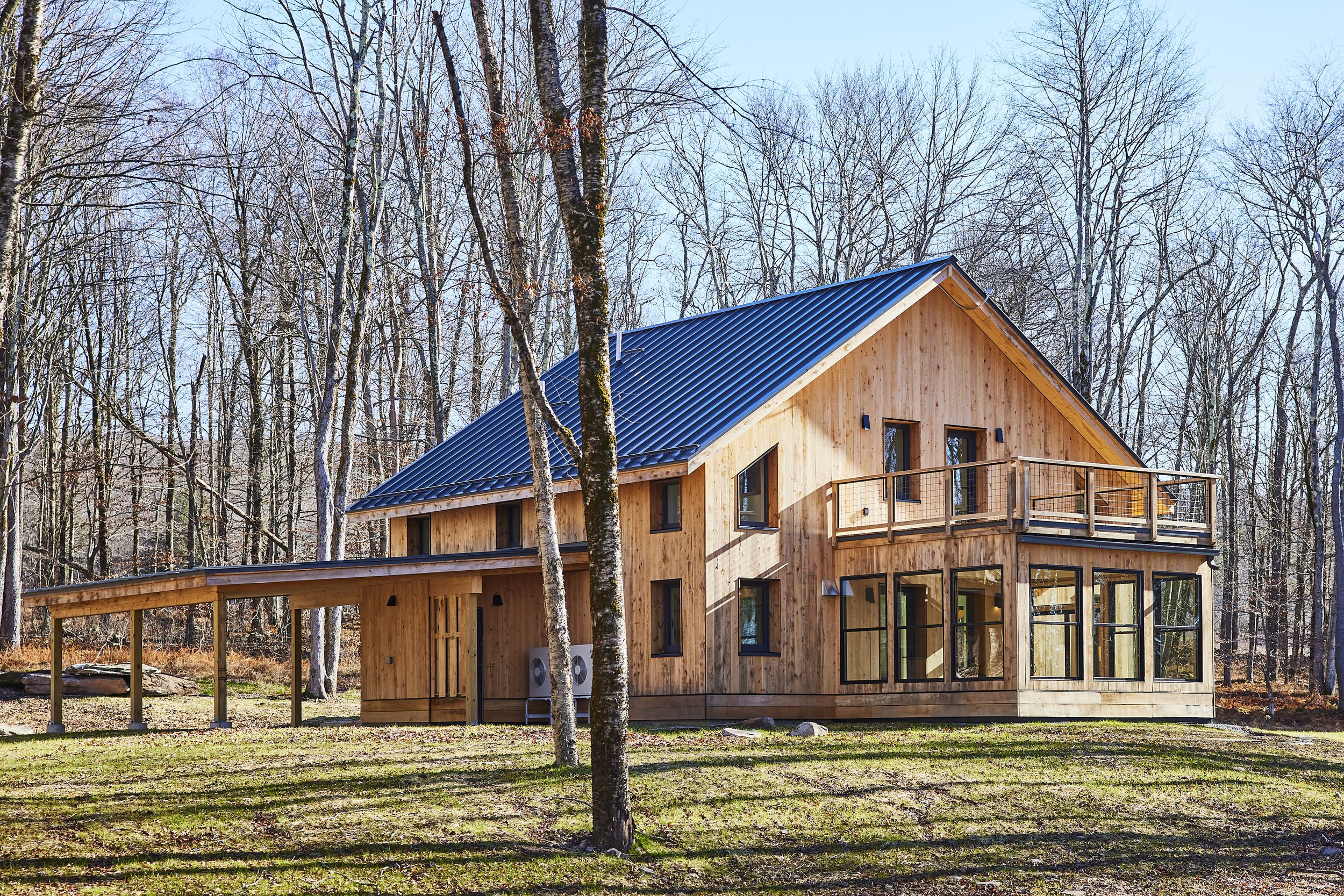 A modern wooden house with large windows and a metal roof in a forested area with leafless trees.