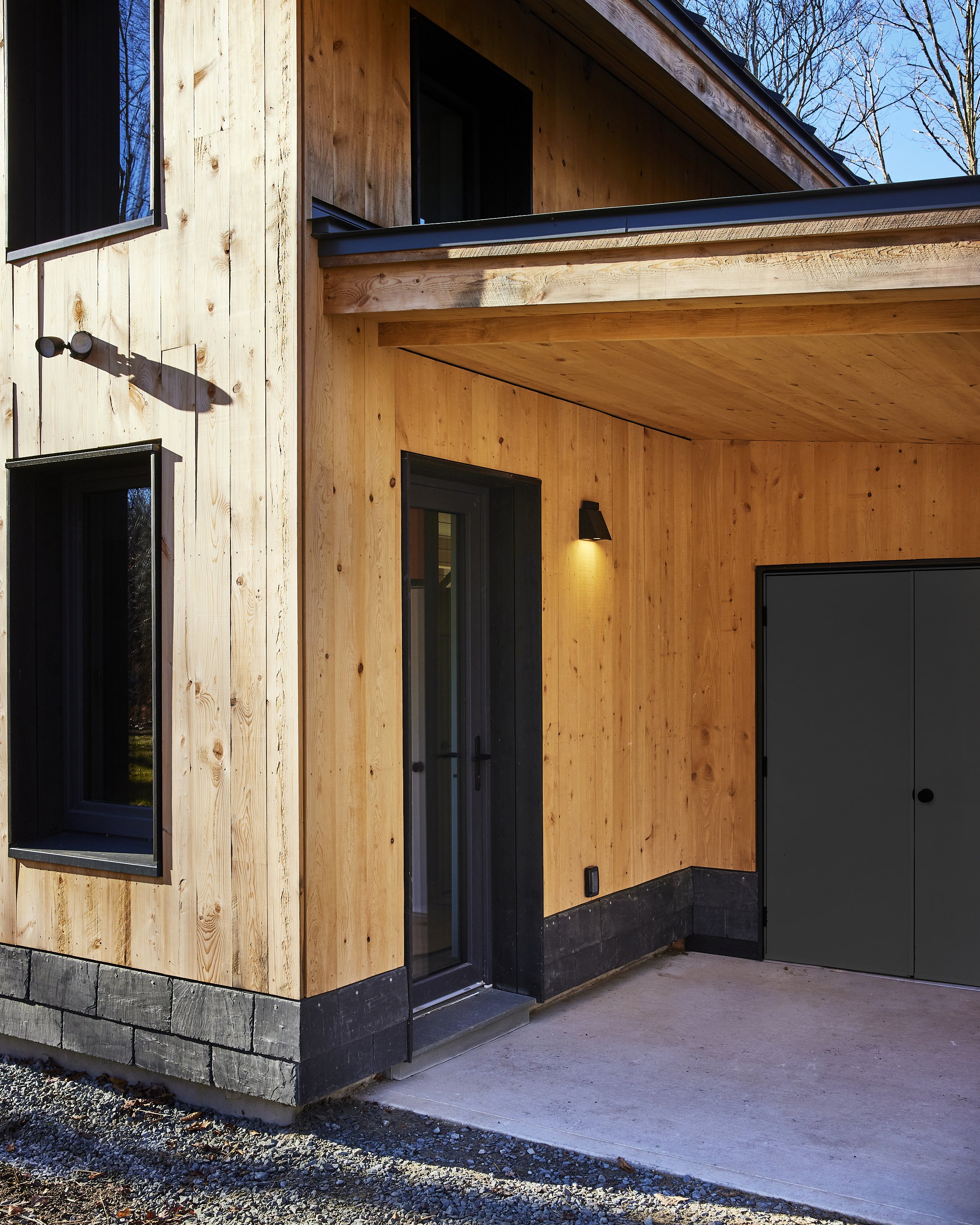Exterior view of a modern house with wood siding, a glass door, and black window frames. There is a small outdoor light fixture and sliding black doors leading to storage or utility space. The house is surrounded by a concrete patio and bare trees in
