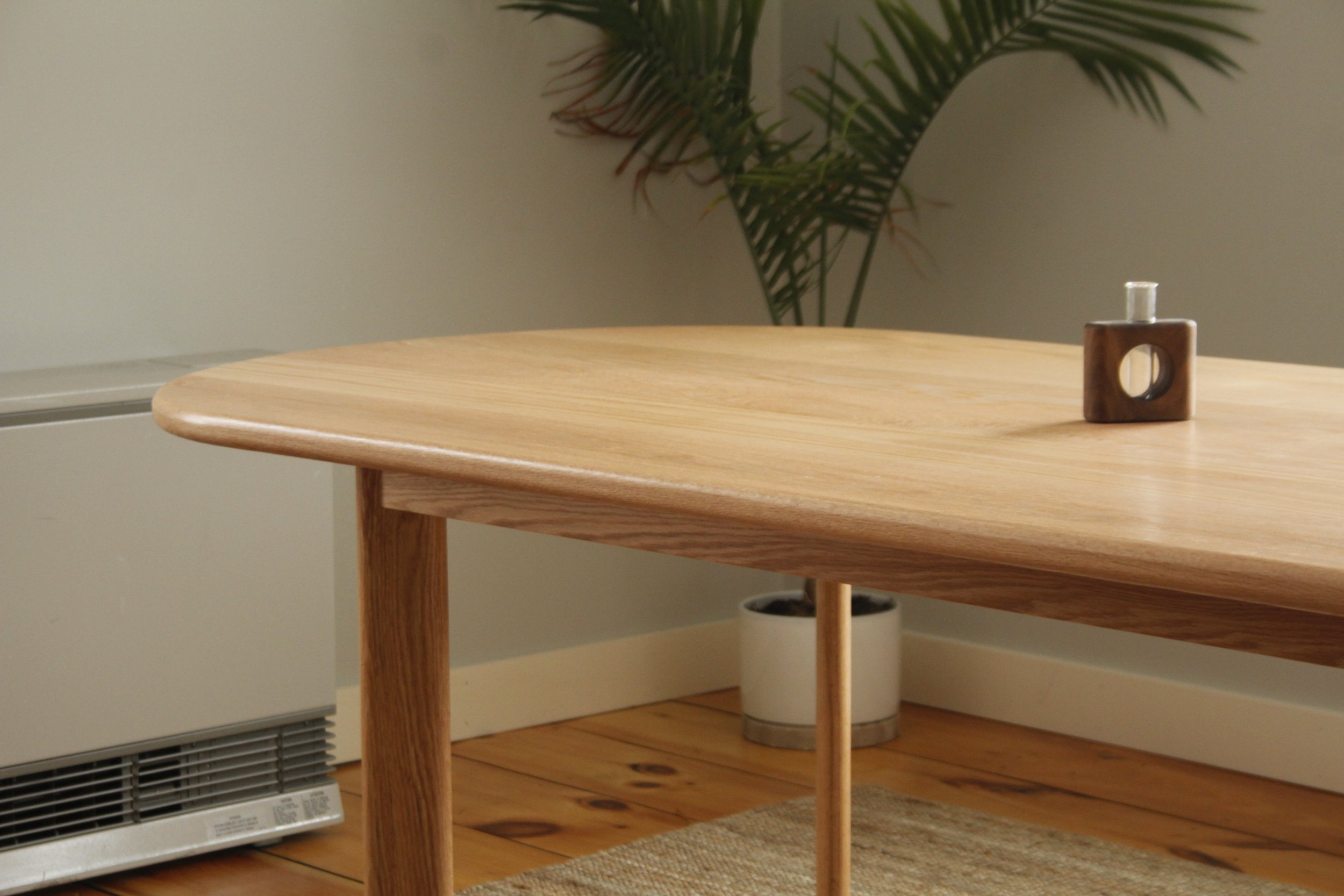 A wooden table with a small square wooden and ceramic object on top, in a room with wooden floor, a potted plant in the background, and a heater along the wall.