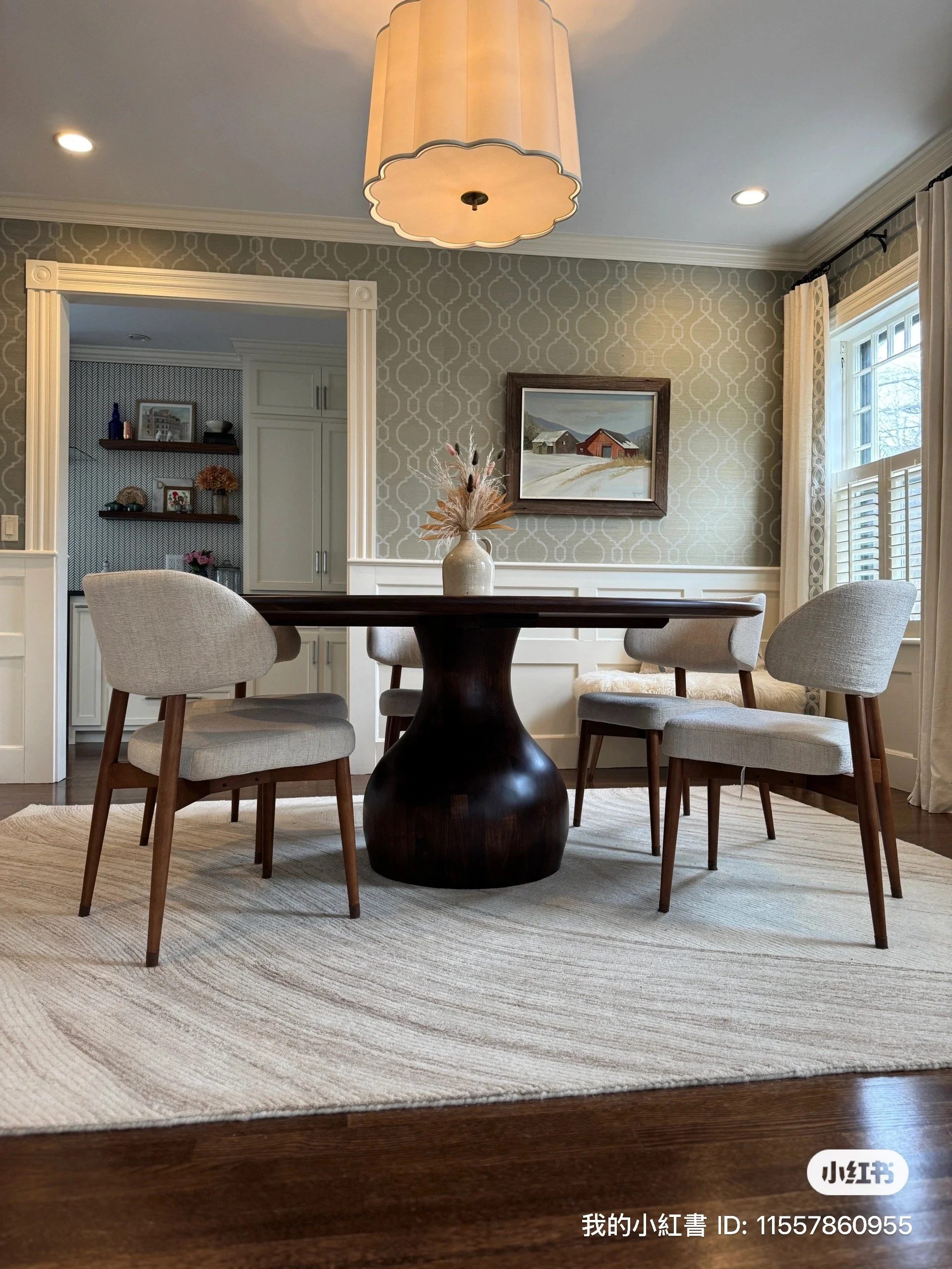 A dining room with a round wooden table, four upholstered chairs, a beige vase with dried flowers as a centerpiece, a patterned wallpapered wall, a framed painting of a farm scene, and a window with curtains letting in natural light.