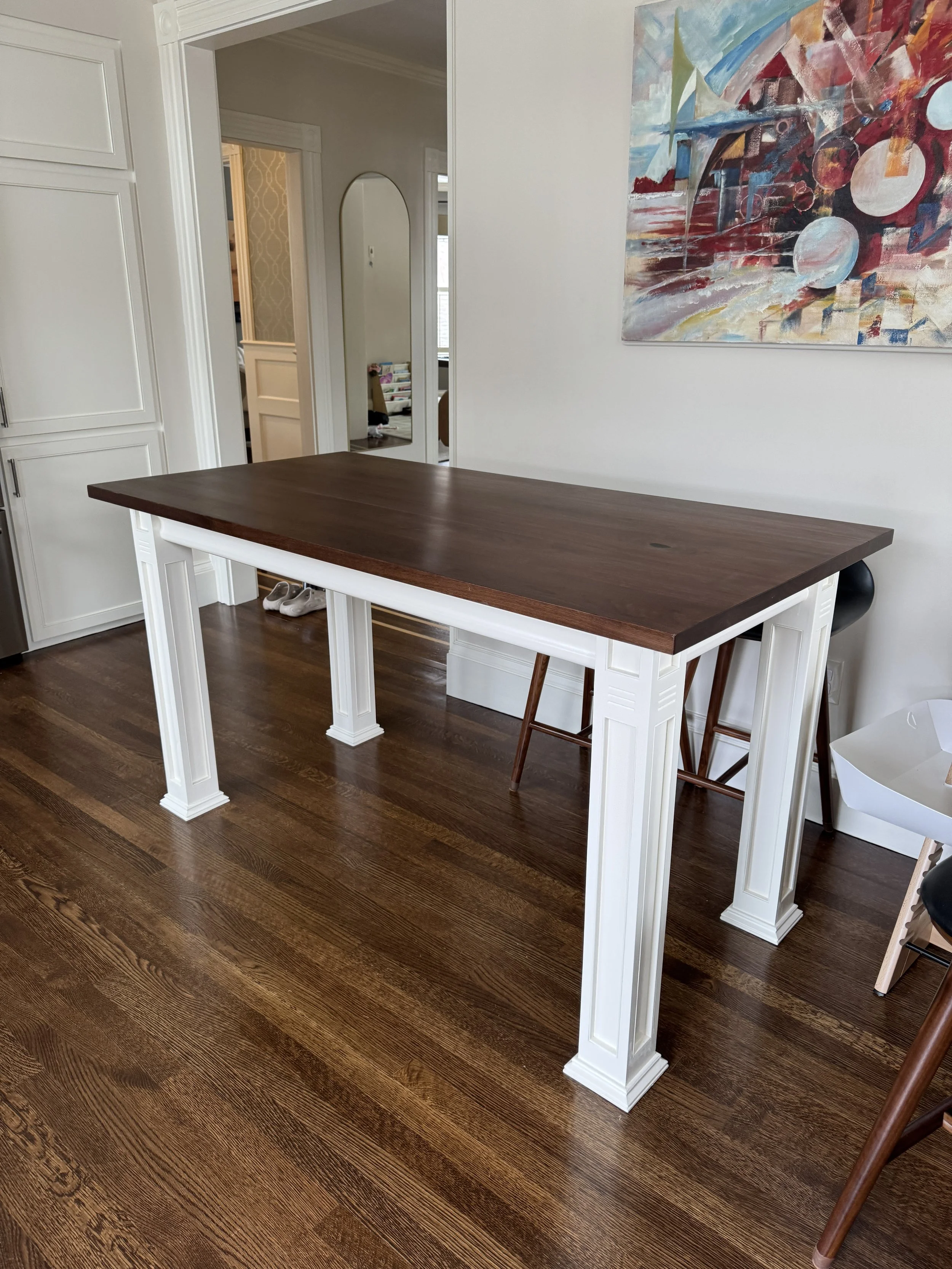 A dining or kitchen table with a dark wood top and white carved legs in a room with hardwood floors. There are chairs and artwork on the walls.