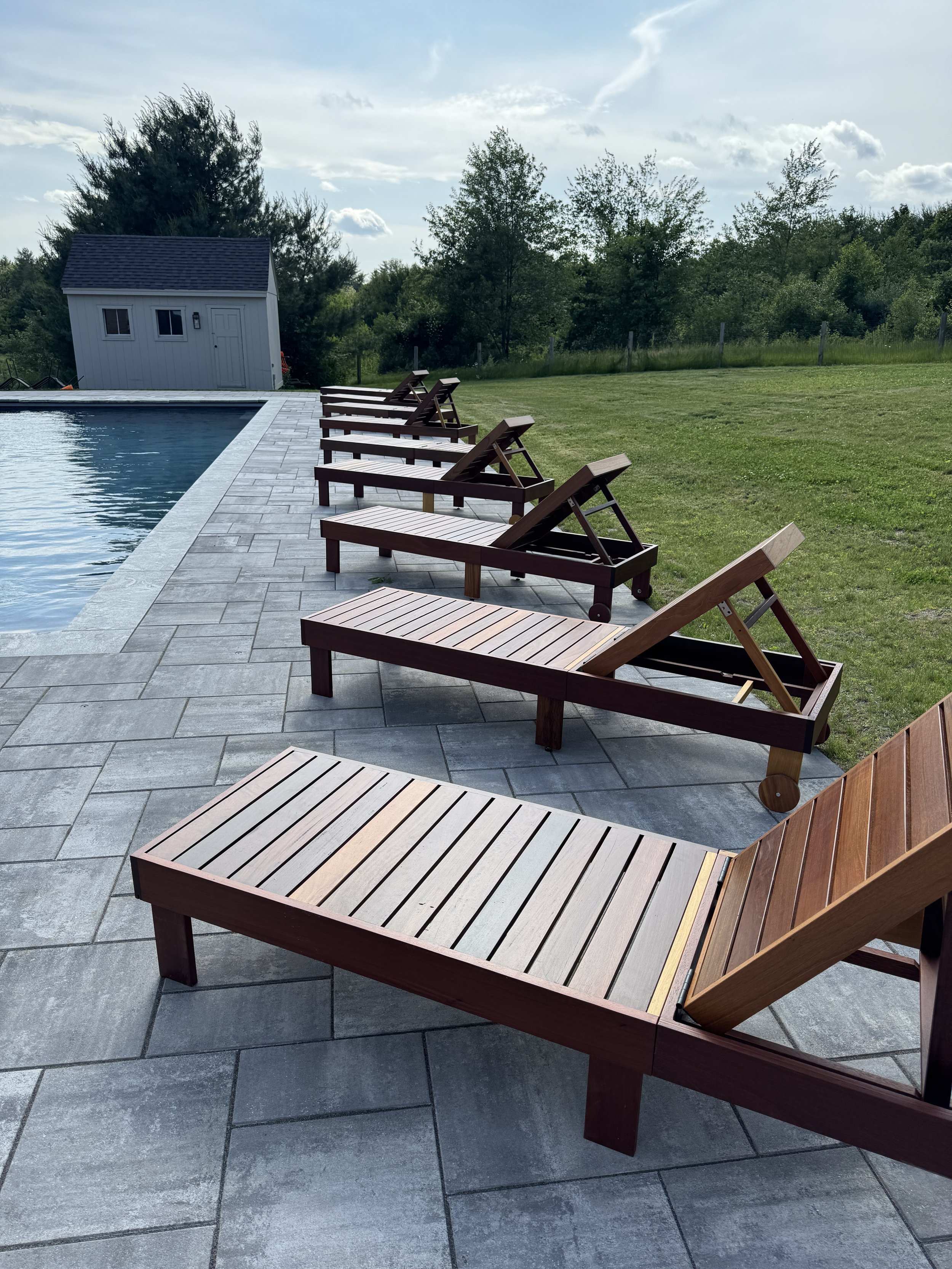 A row of wooden lounge chairs by a swimming pool on a grey tiled pool deck, with a green grassy yard and trees in the background under a partly cloudy sky.