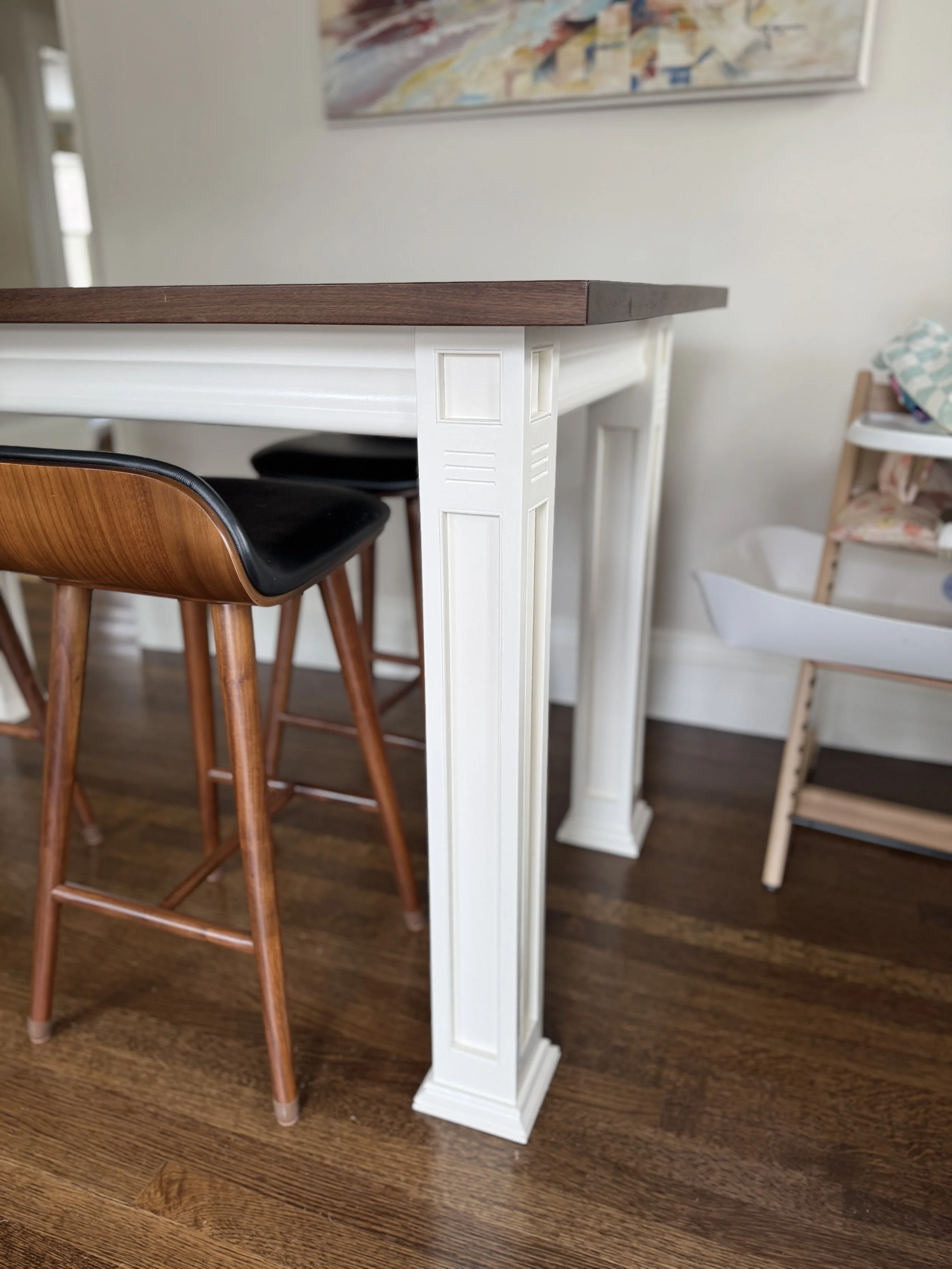 Close-up of a kitchen or bar-height table with ornate white legs and a dark wooden top, with a wooden chair nearby and a play area in the background.